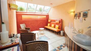 A sunlit room with a red wall, a wooden bench with yellow cushions, wicker chairs, a small round table, and a large sink.