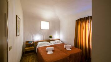 Habitación de hotel con una cama individual, una mesita de noche con lámpara, un ventanal pequeño y una cortina.