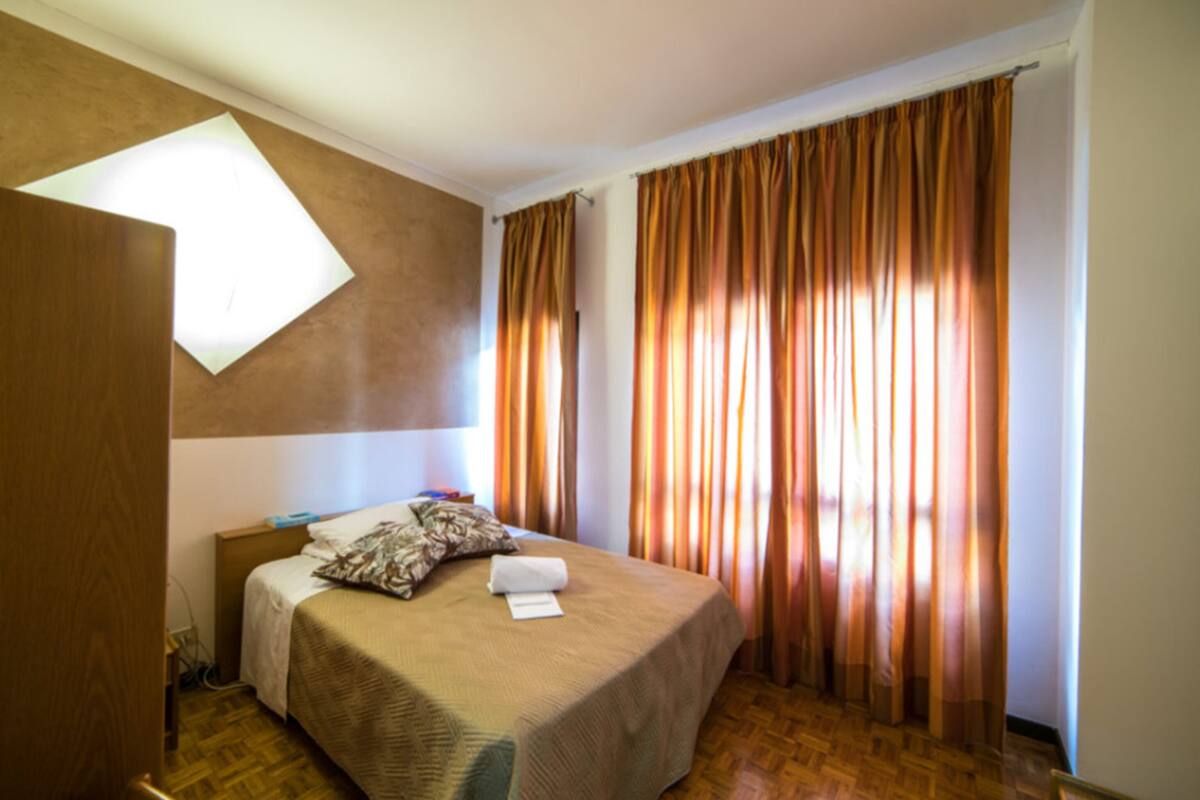 A hotel room with a bed, a nightstand, a window with curtains, and a wall-mounted light fixture.