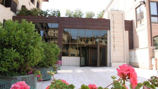 A hotel entrance with a glass facade, a brown awning, and a garden with pink flowers. - Hotel La Vecchia Cartiera (Colle di Val d'Elsa)
