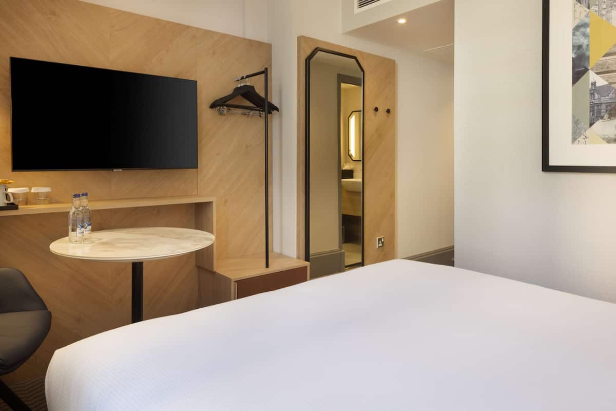 A hotel room with a bed, a TV mounted on the wall, a small round table with a bottle and cups, a chair, and a mirror.