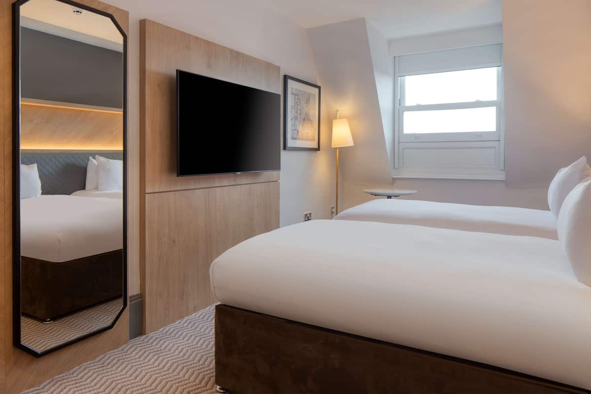 A hotel room with two beds, a flat-screen TV, a large mirror, and a window with natural light.