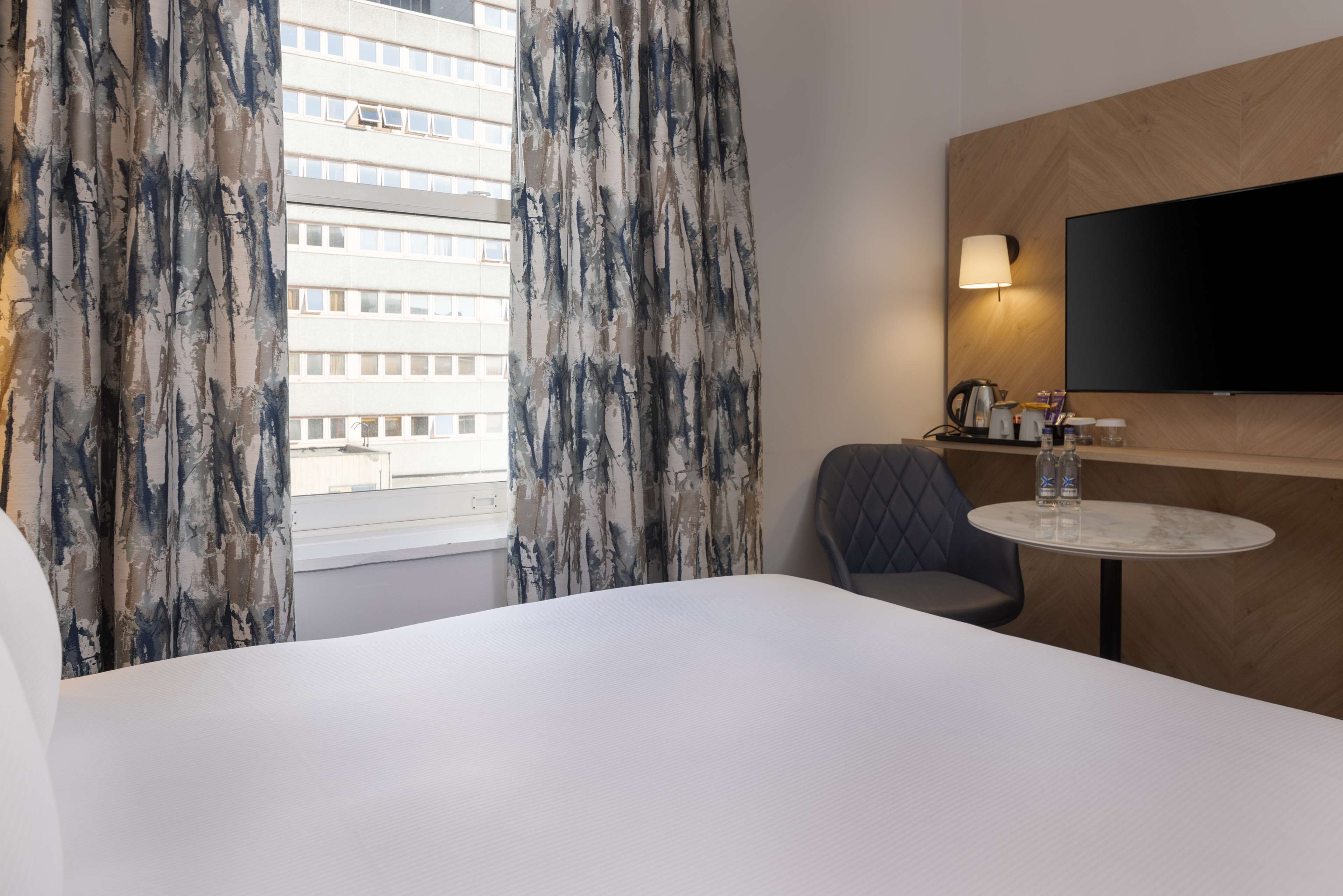 A hotel room with a bed, a round table with a chair, a flat-screen TV mounted on the wall, and a window with curtains.