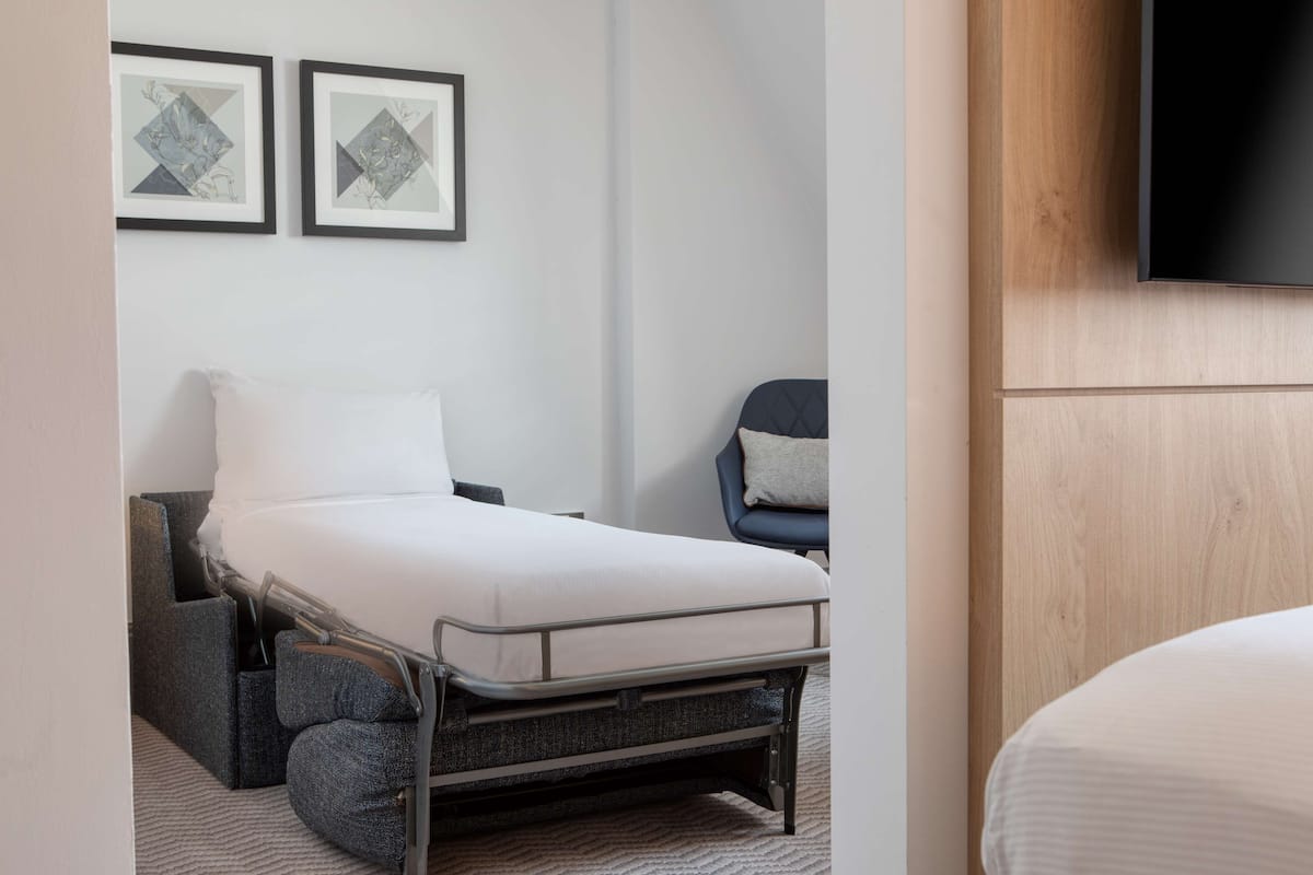 A compact hotel room with a bed, a chair, and two framed artworks on the wall.
