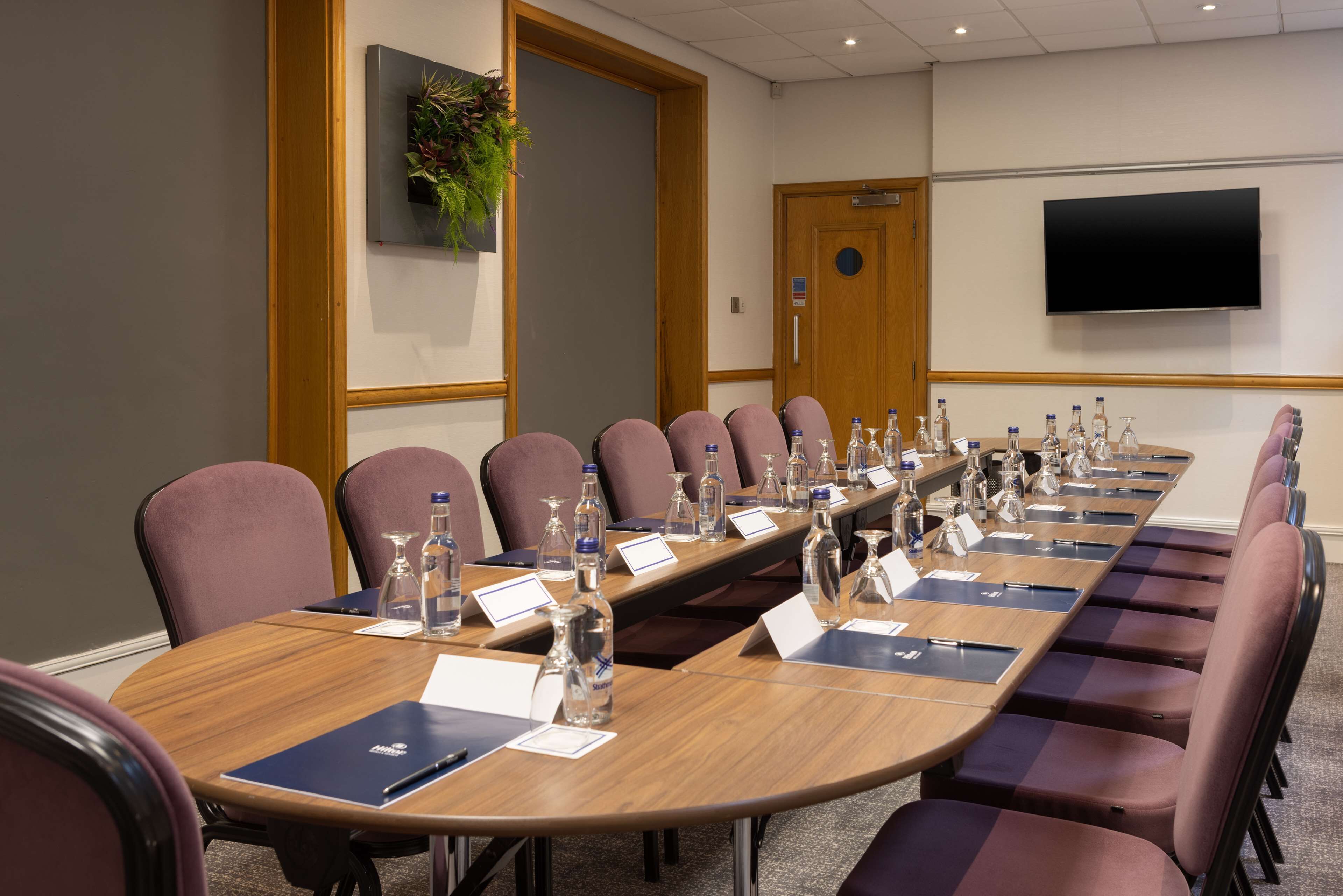 A conference room with a long table, chairs, nameplates, water bottles, and a TV on the wall.