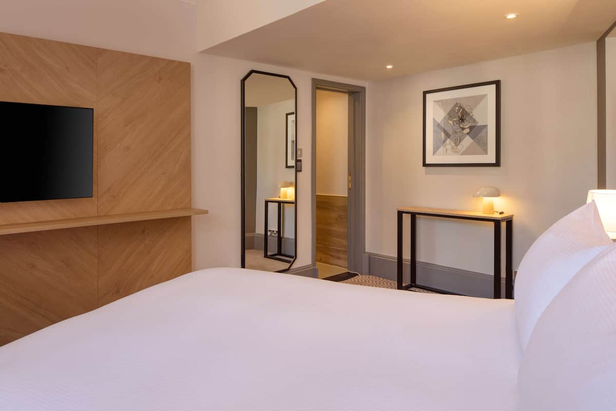 A hotel room with a large bed, a mounted TV, a bedside table with a lamp, a framed artwork on the wall, and a mirror reflecting the room.