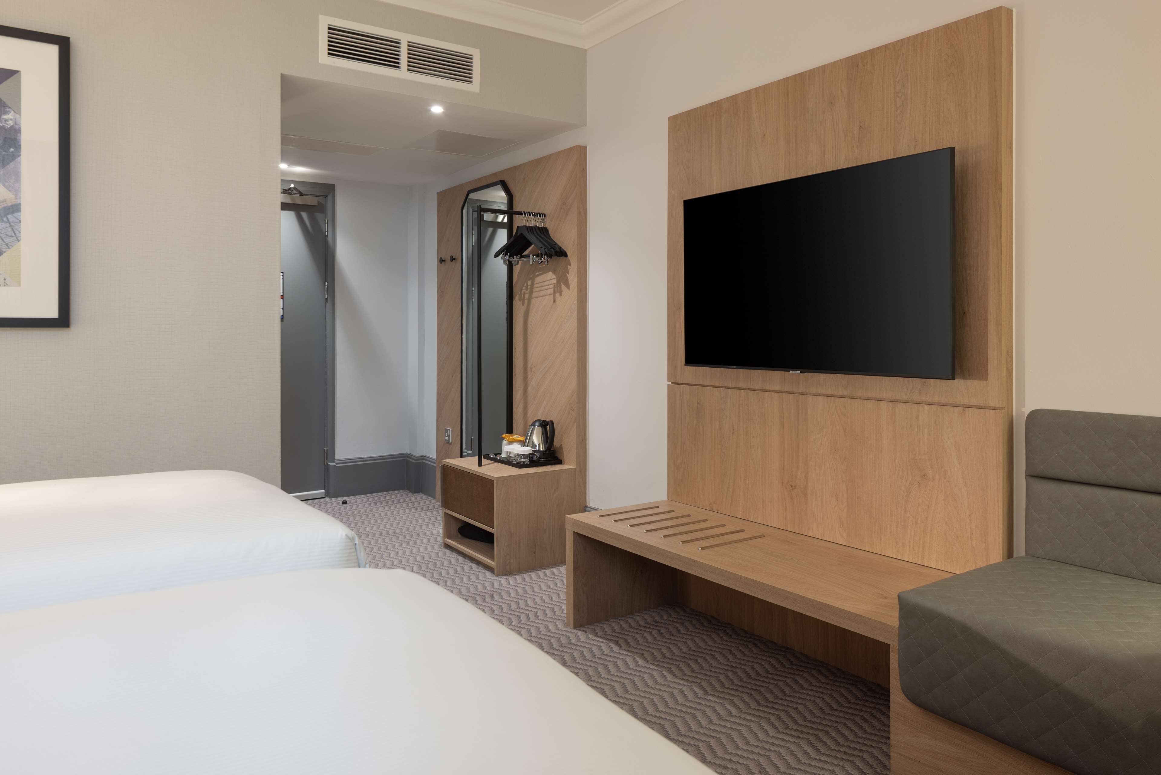 A hotel room with two beds, a TV mounted on the wall, a bench, a mirror, and a coat rack.