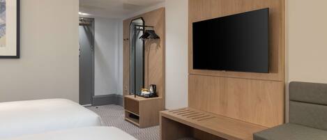 A hotel room with two beds, a TV mounted on the wall, a bench, a mirror, and a coat rack.