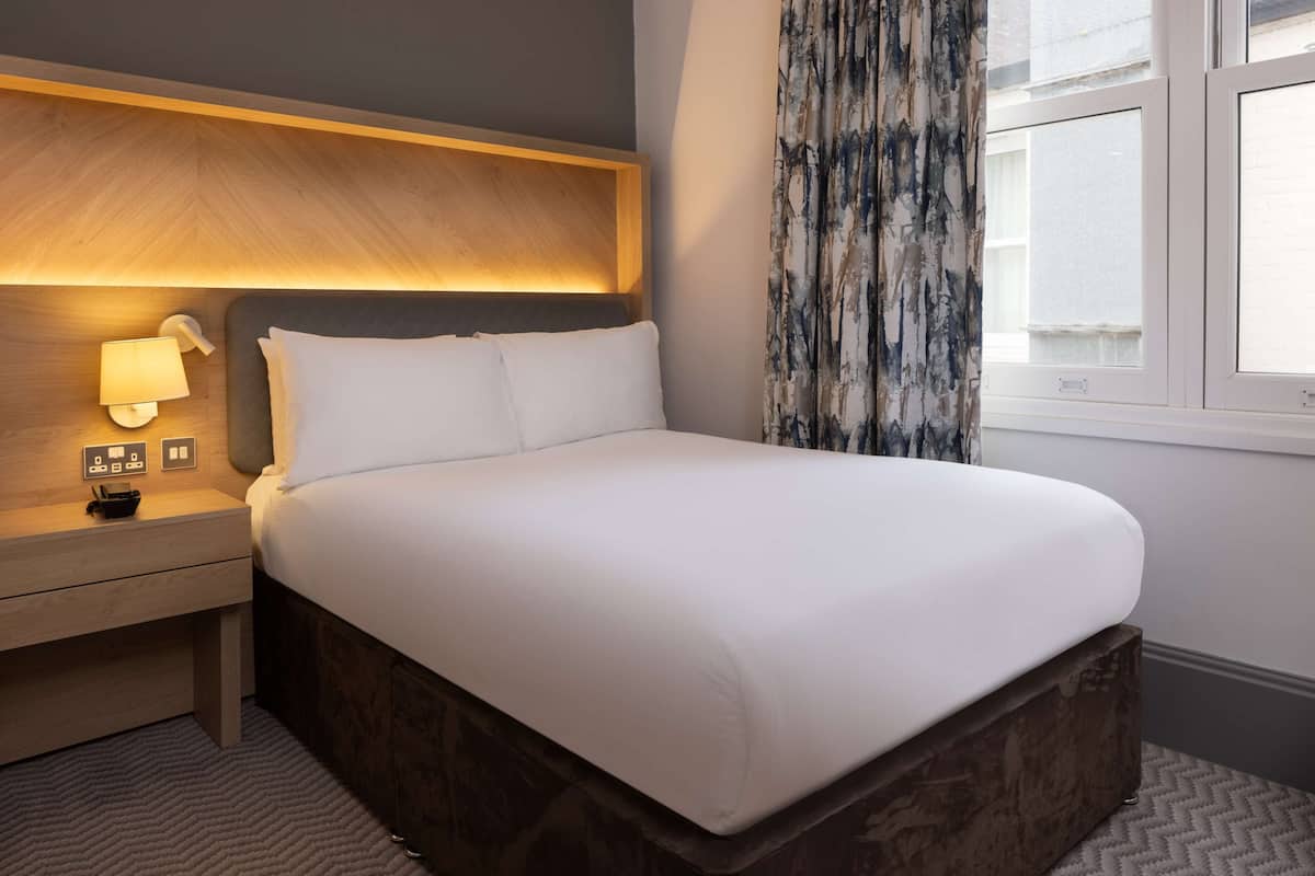 A hotel room with a bed, bedside table, lamp, and a window with curtains.