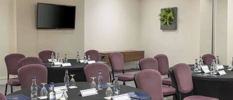 A conference room with round tables, chairs, nameplates, water bottles, and a flat-screen TV on the wall.