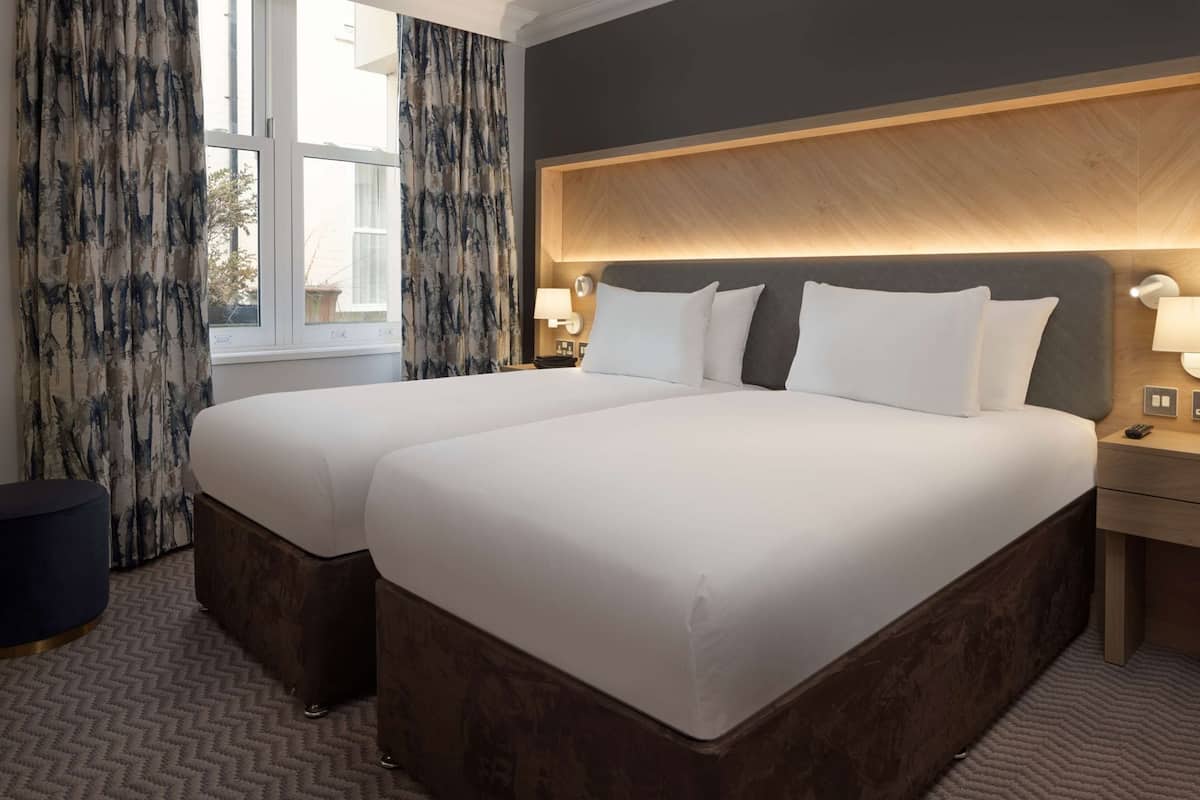 A double bed with a wooden headboard, a bedside table with a lamp, and a view of the outdoors through a window with curtains.