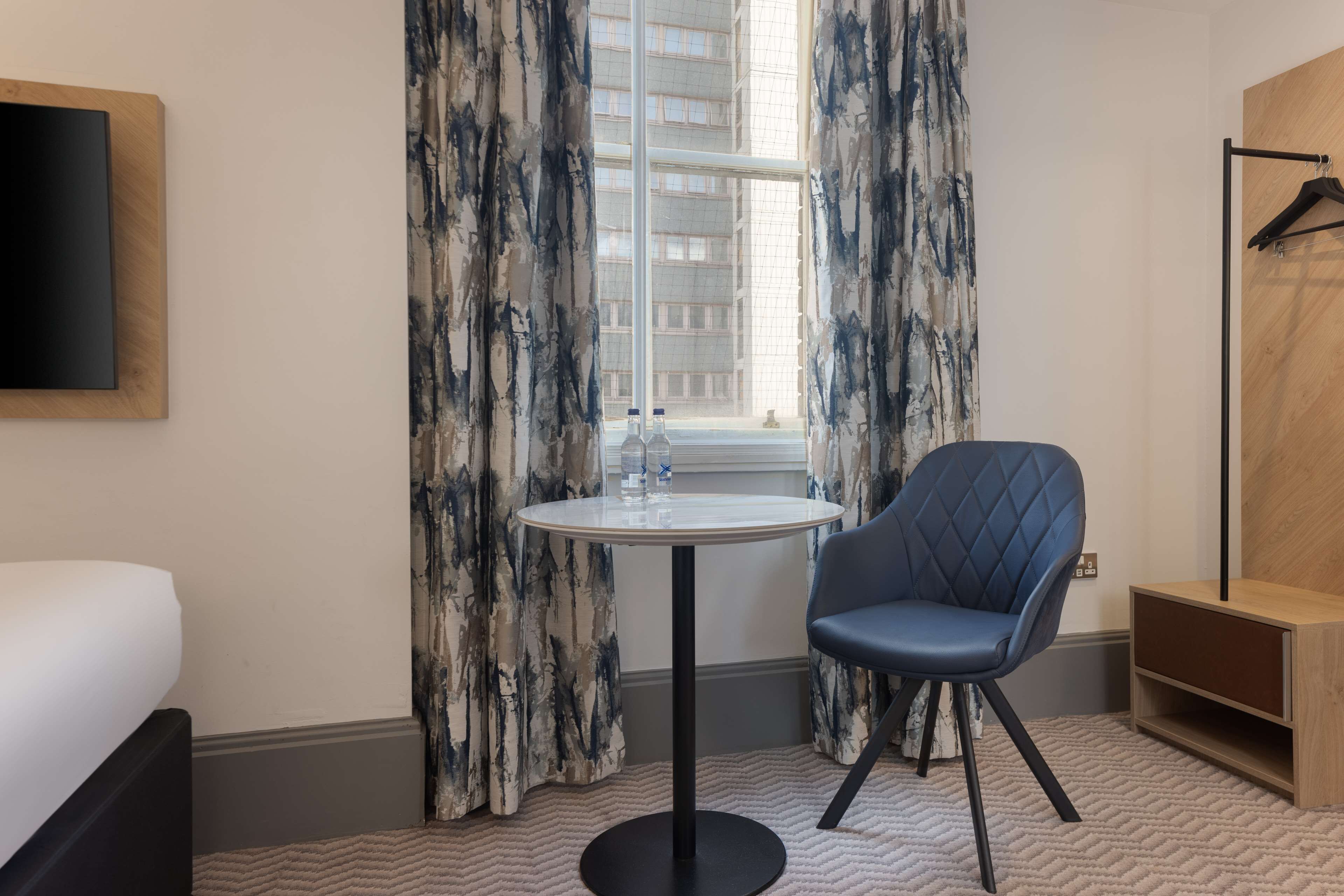 A hotel room with a bed, a round table with a sink, a blue chair, and a wooden cabinet.