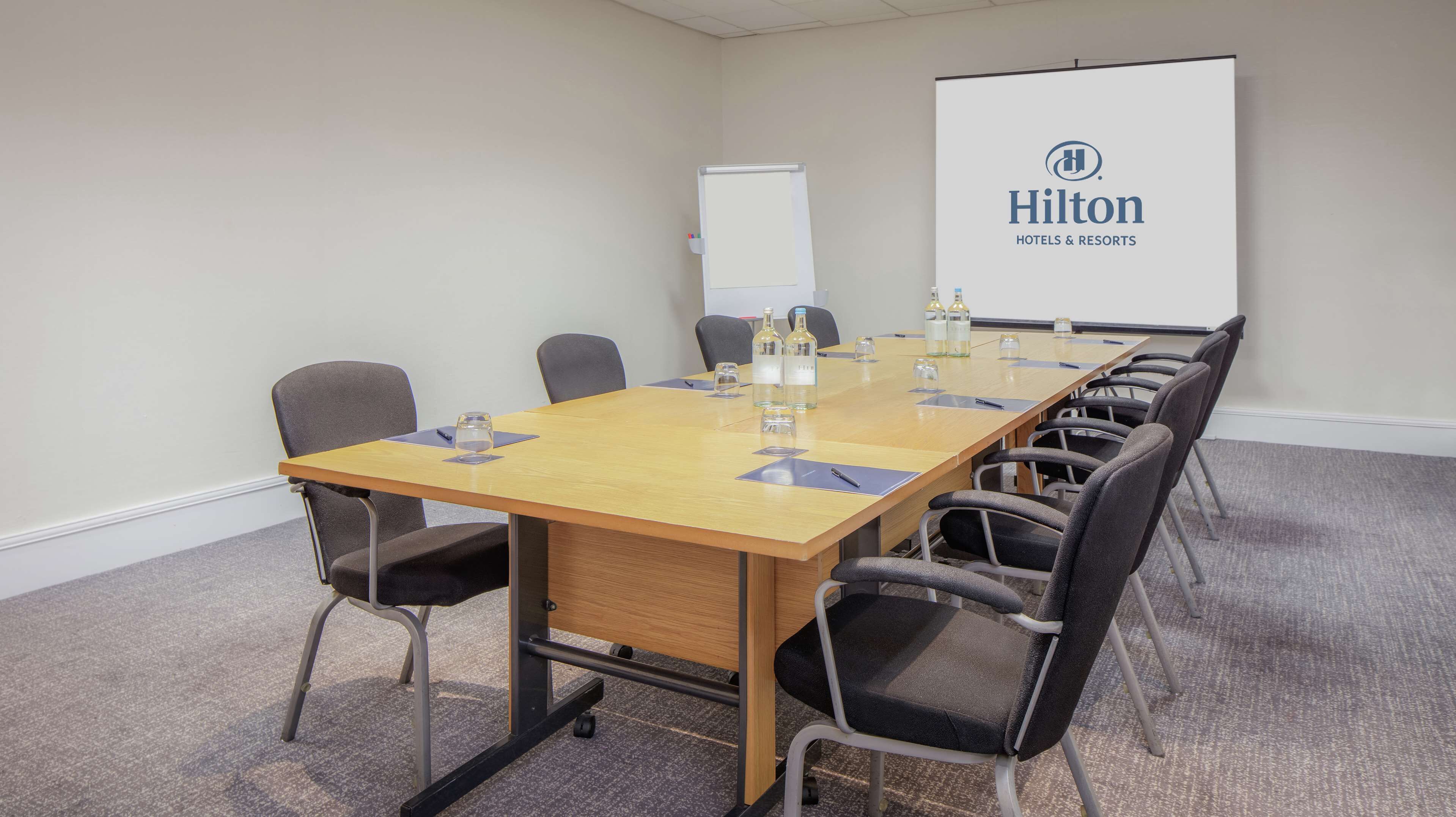 Una sala de conferencias con una larga mesa, sillas y una pantalla proyectora mostrando el logotipo de Hilton Hotels & Resorts.