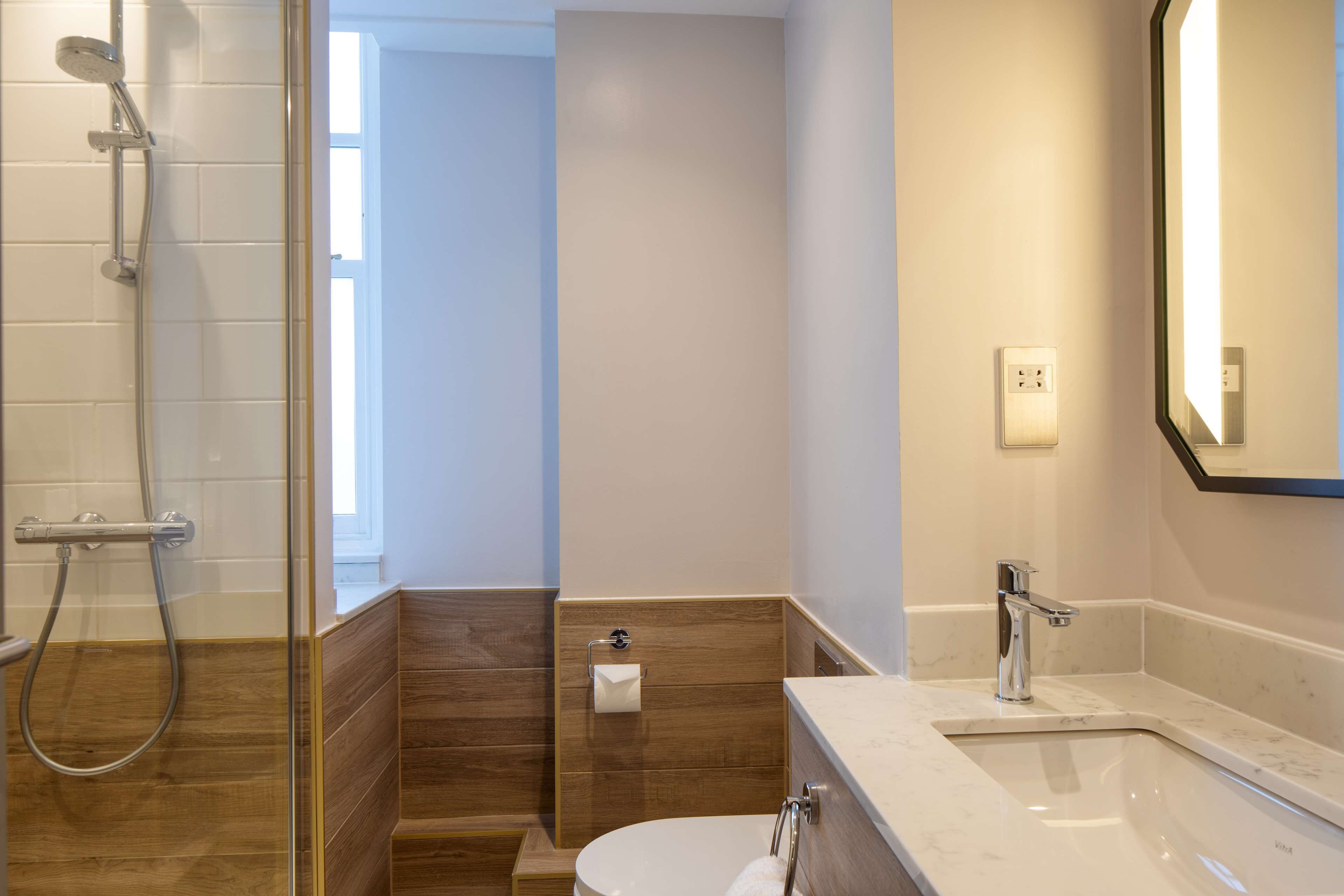 A modern bathroom with a glass shower enclosure, a wooden vanity, a white toilet, and a large mirror.
