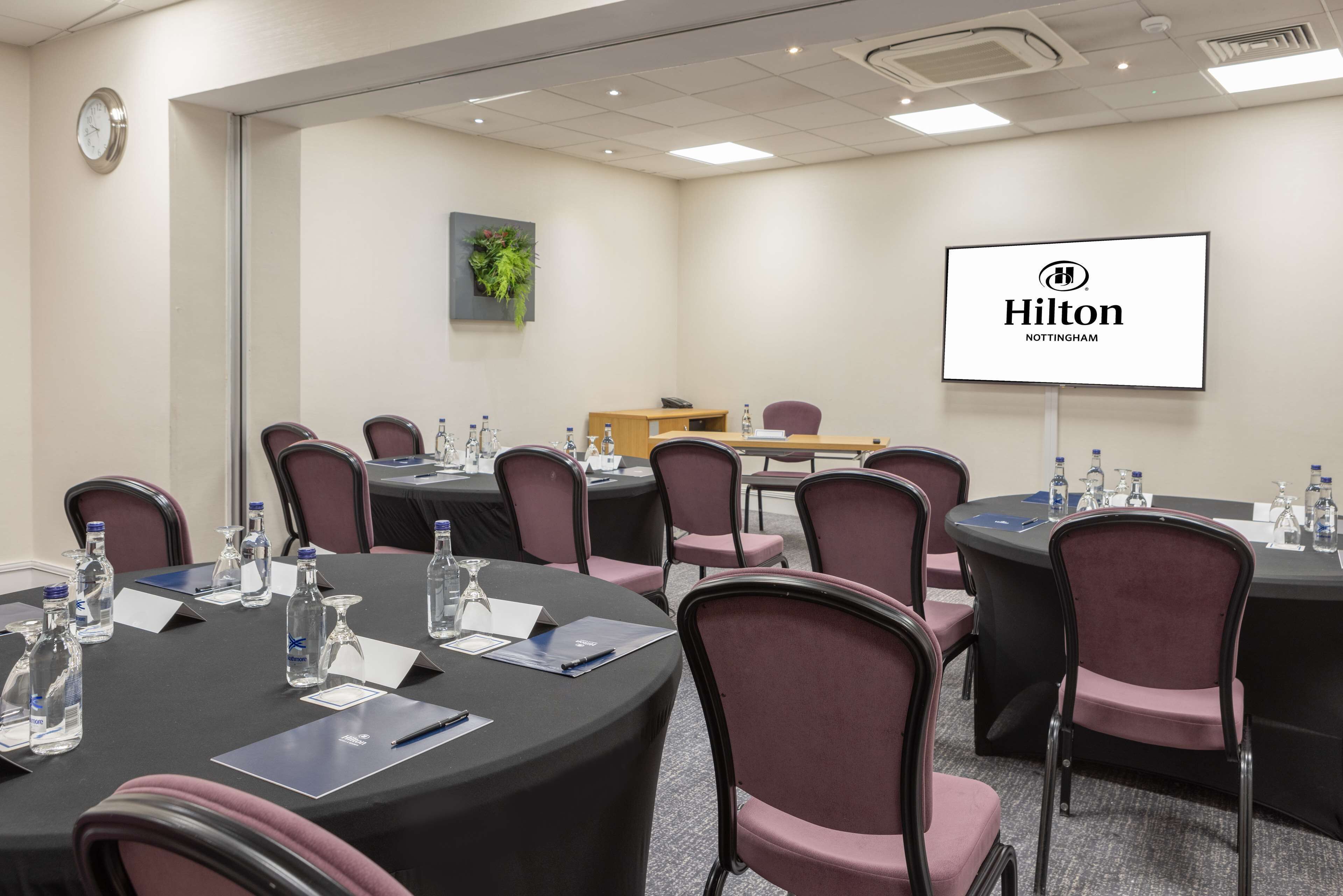 A conference room with round tables, chairs, water bottles, and a screen displaying the Hilton logo.
