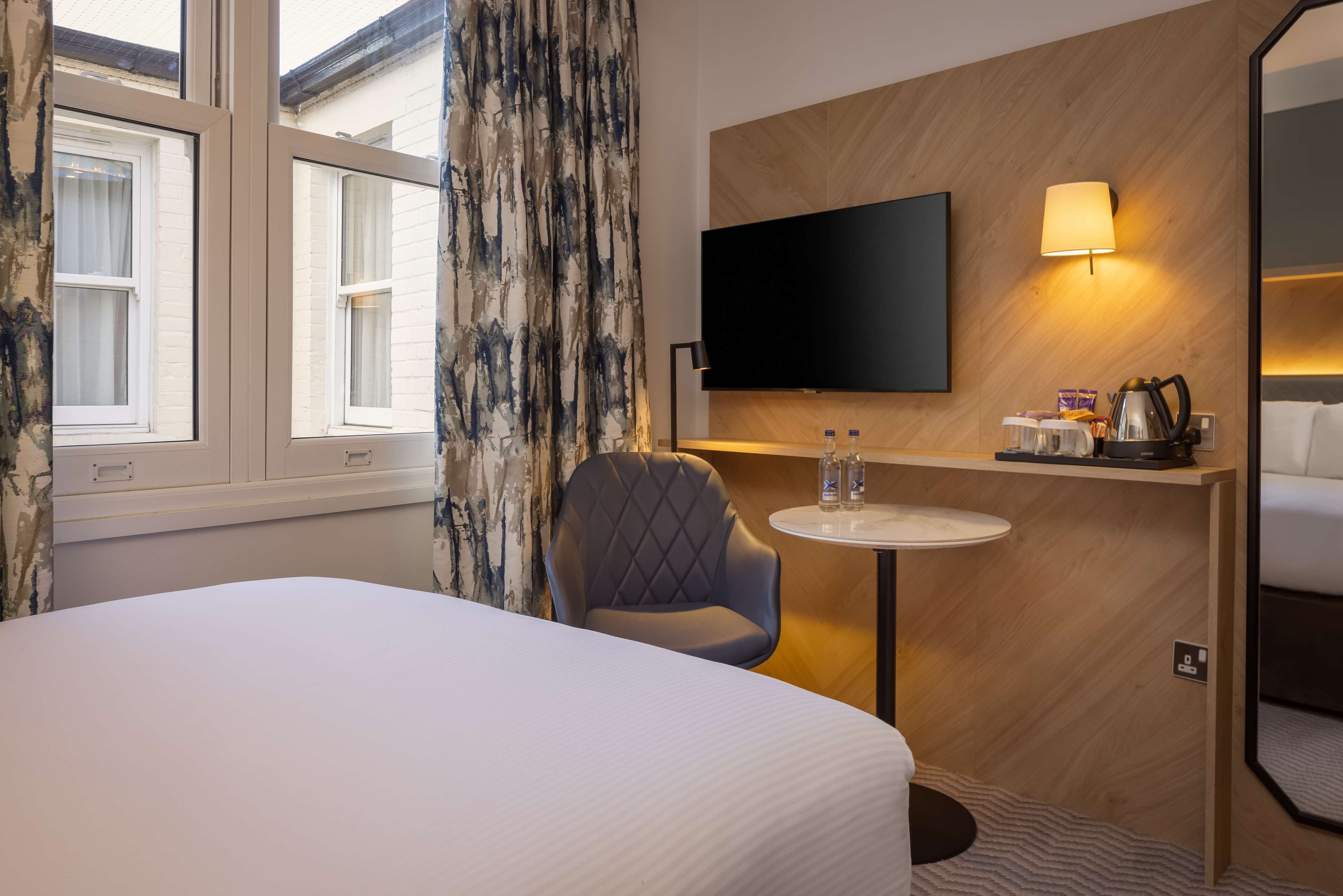 A hotel room with a bed, a chair, a small table with a lamp, a TV, and a window with curtains.