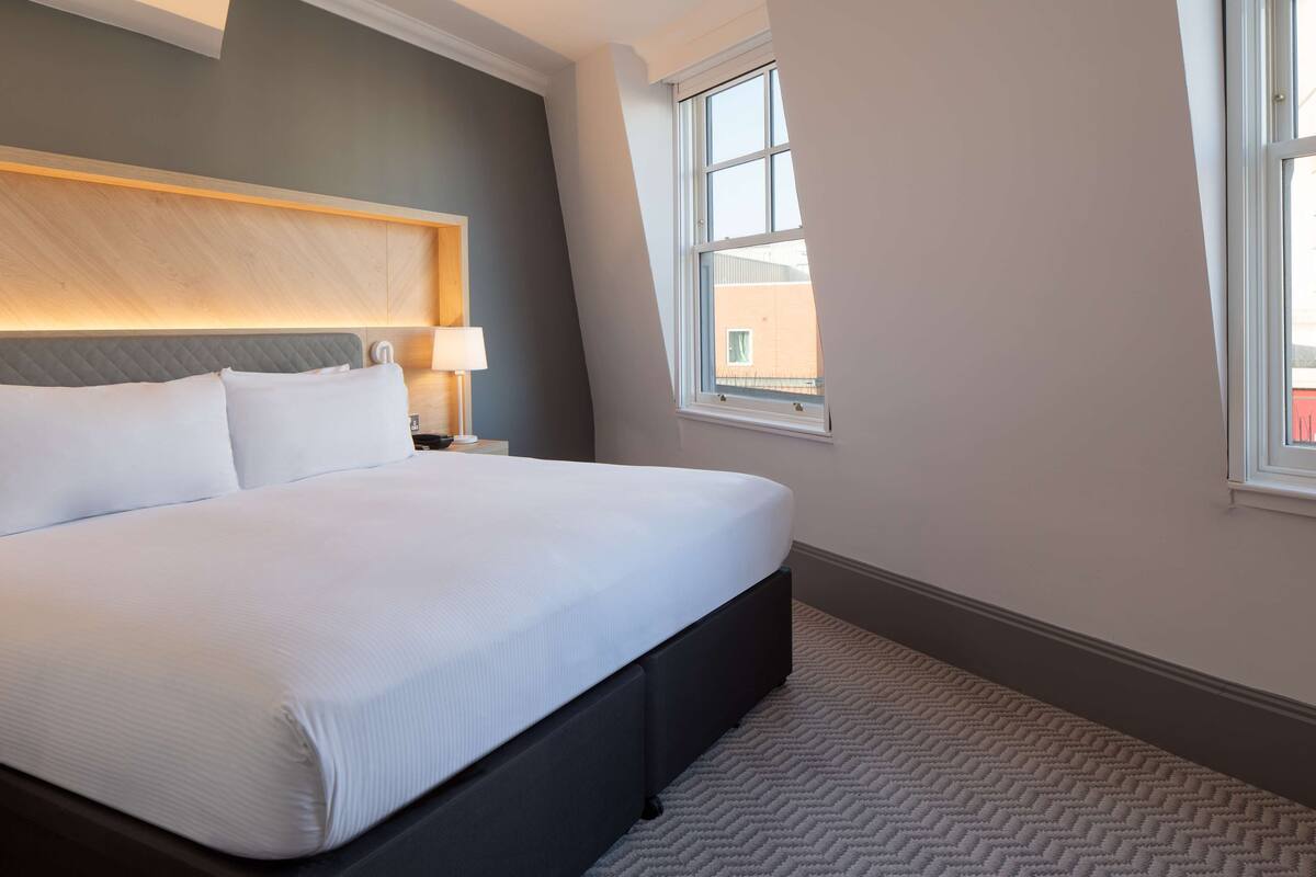 A neatly made bed in a hotel room with a headboard, a nightstand, a lamp, and a window with a view of buildings outside.
