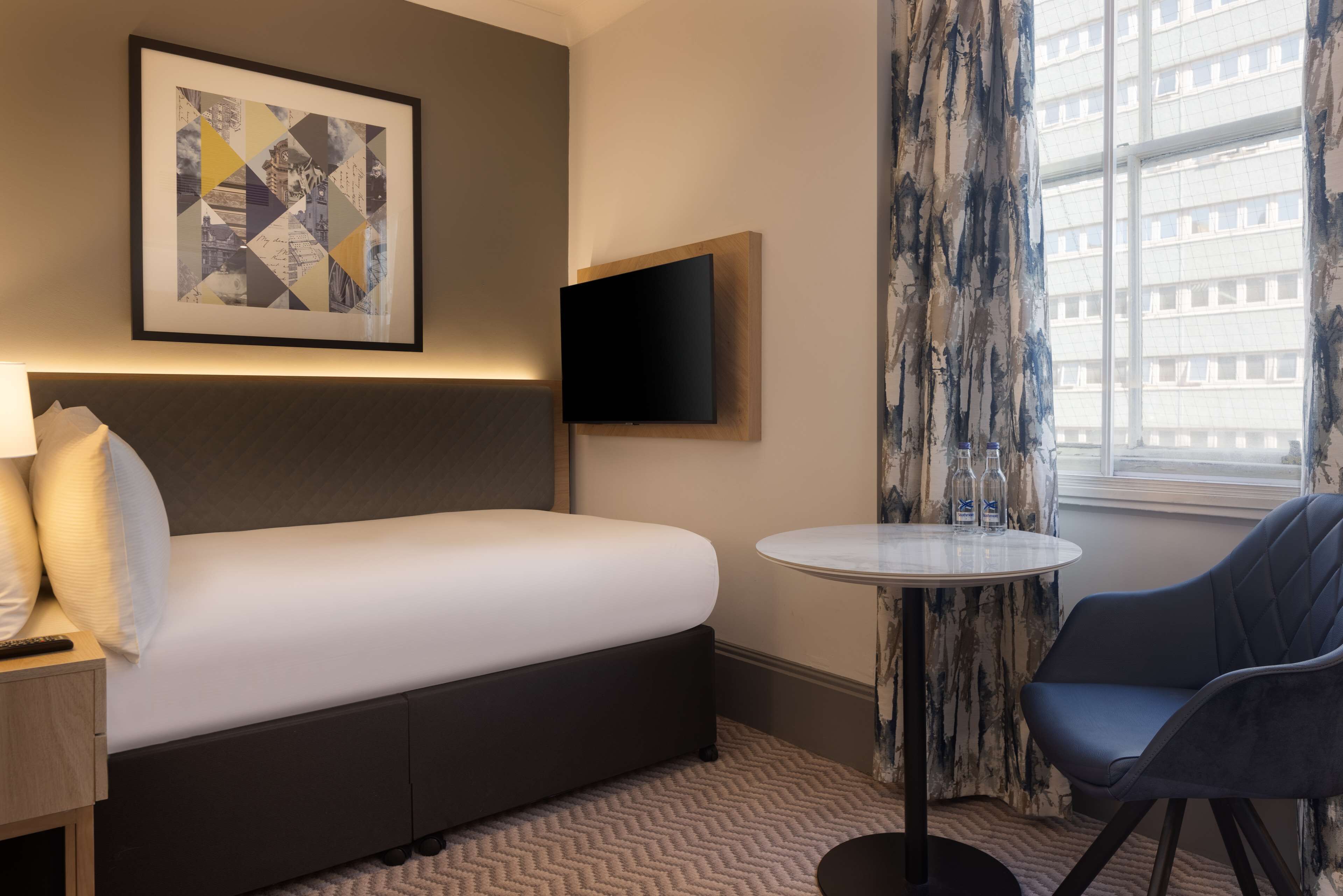 A hotel room with a bed, a TV mounted on the wall, a small round table, and a chair.
