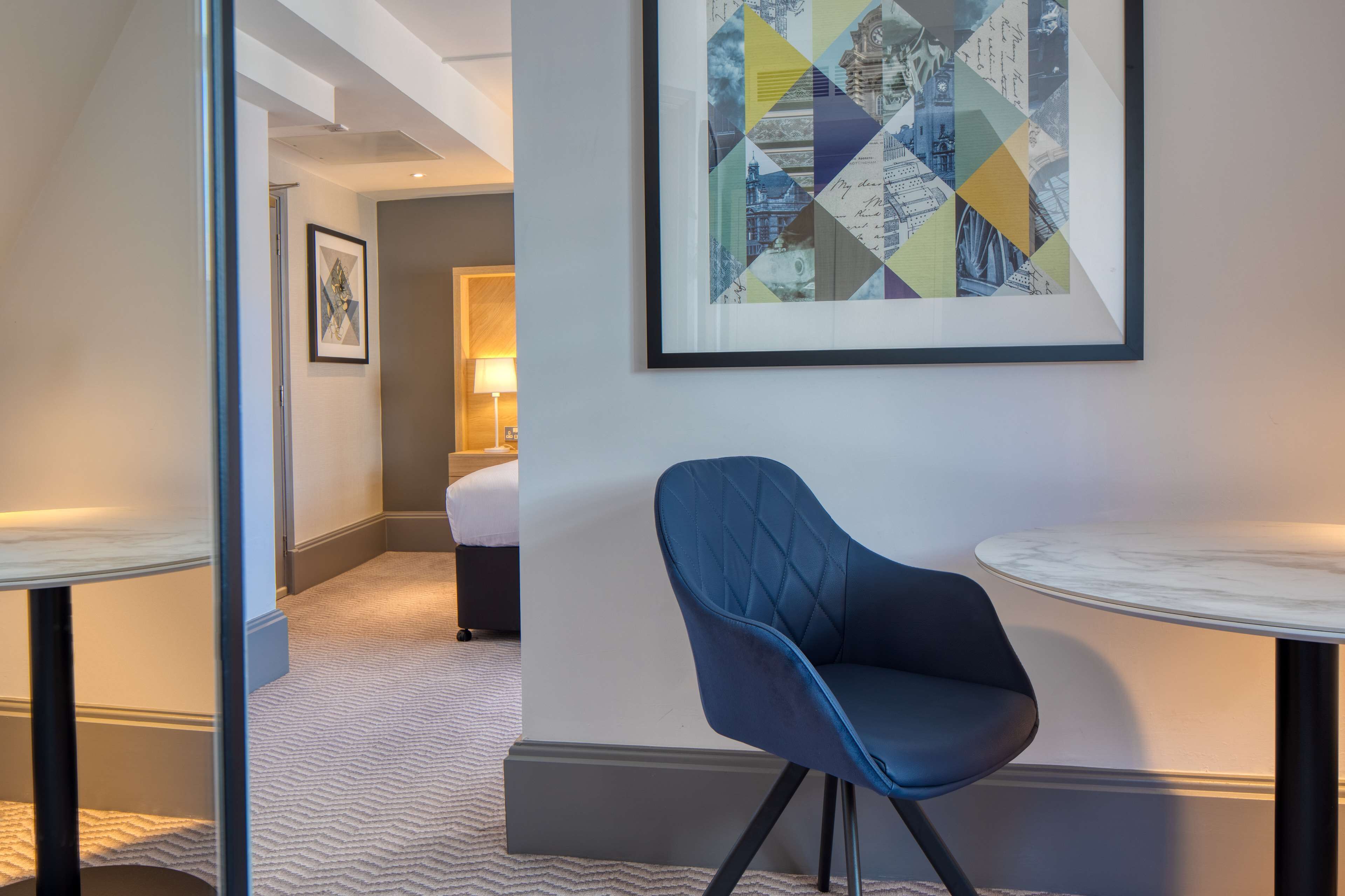Une chambre d’hôtel avec une chaise bleue, une table ronde et une œuvre d’art encadrée accrochée au mur.
