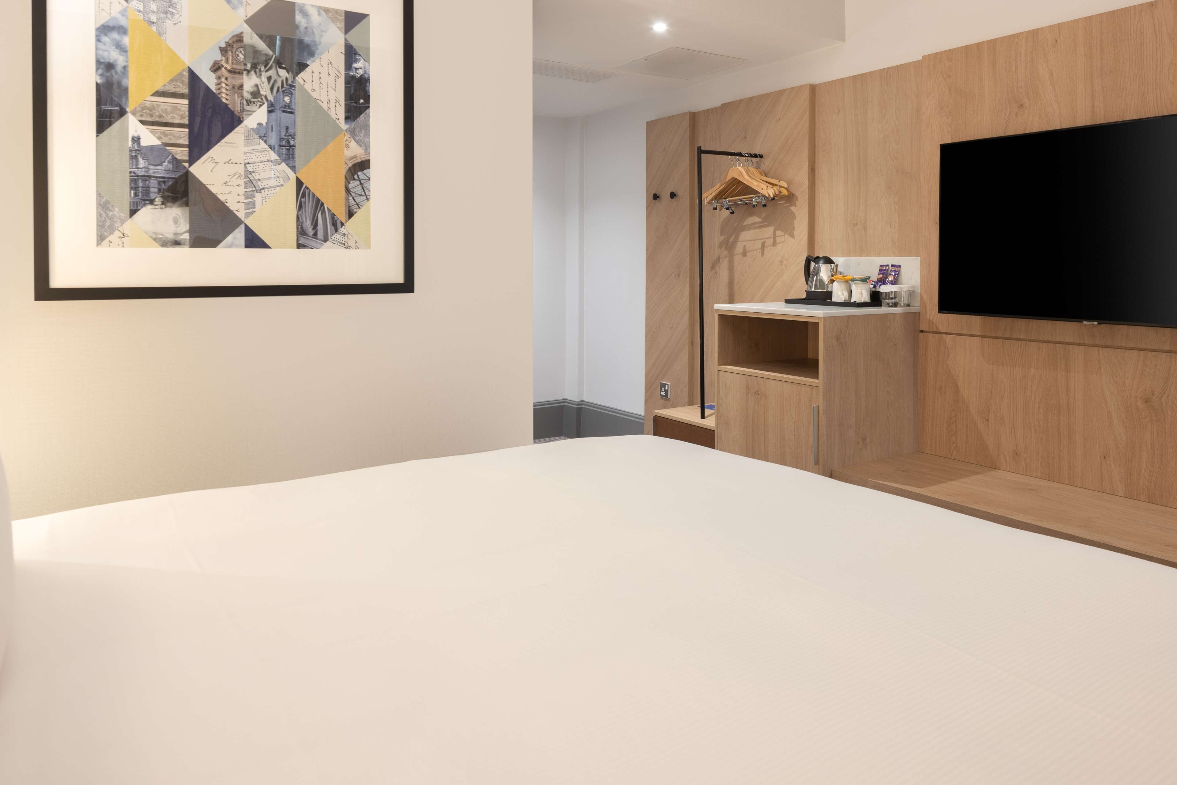 A modern hotel room with a large bed, a mounted TV, wooden paneling, and a framed artwork on the wall.