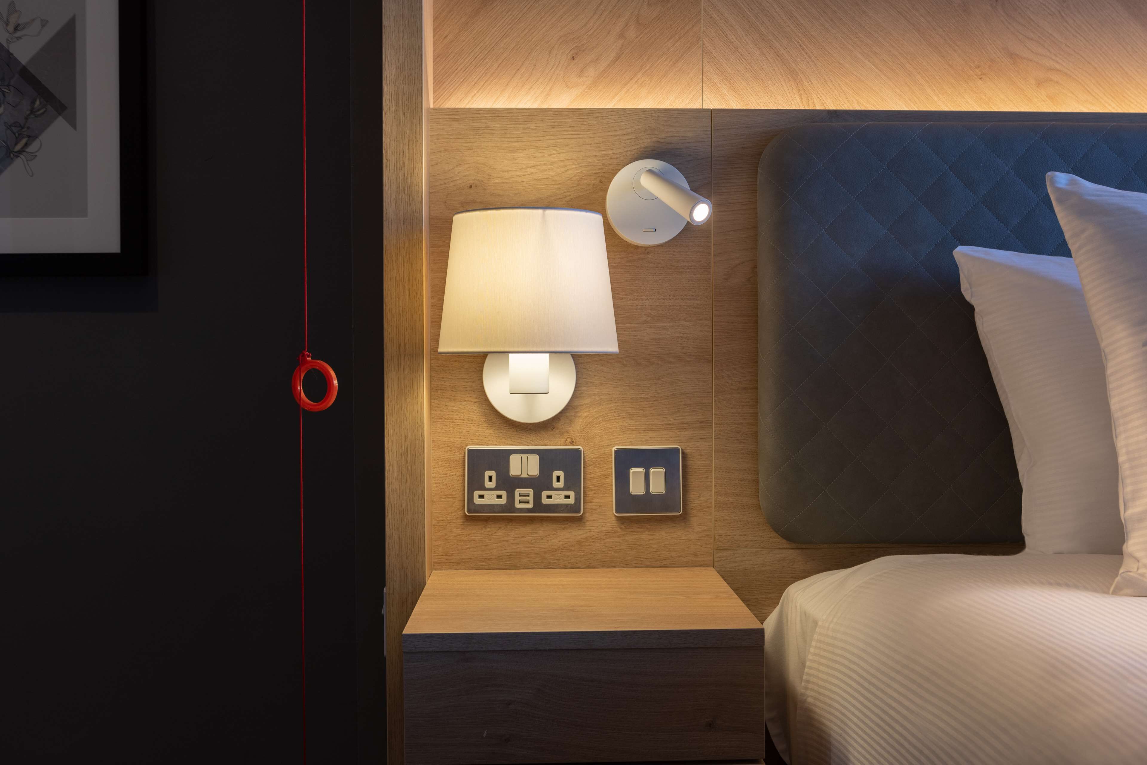 Une chambre d’hôtel bien rangée, avec un lit, une table de chevet, une lampe fixée au mur et une prise de courant.