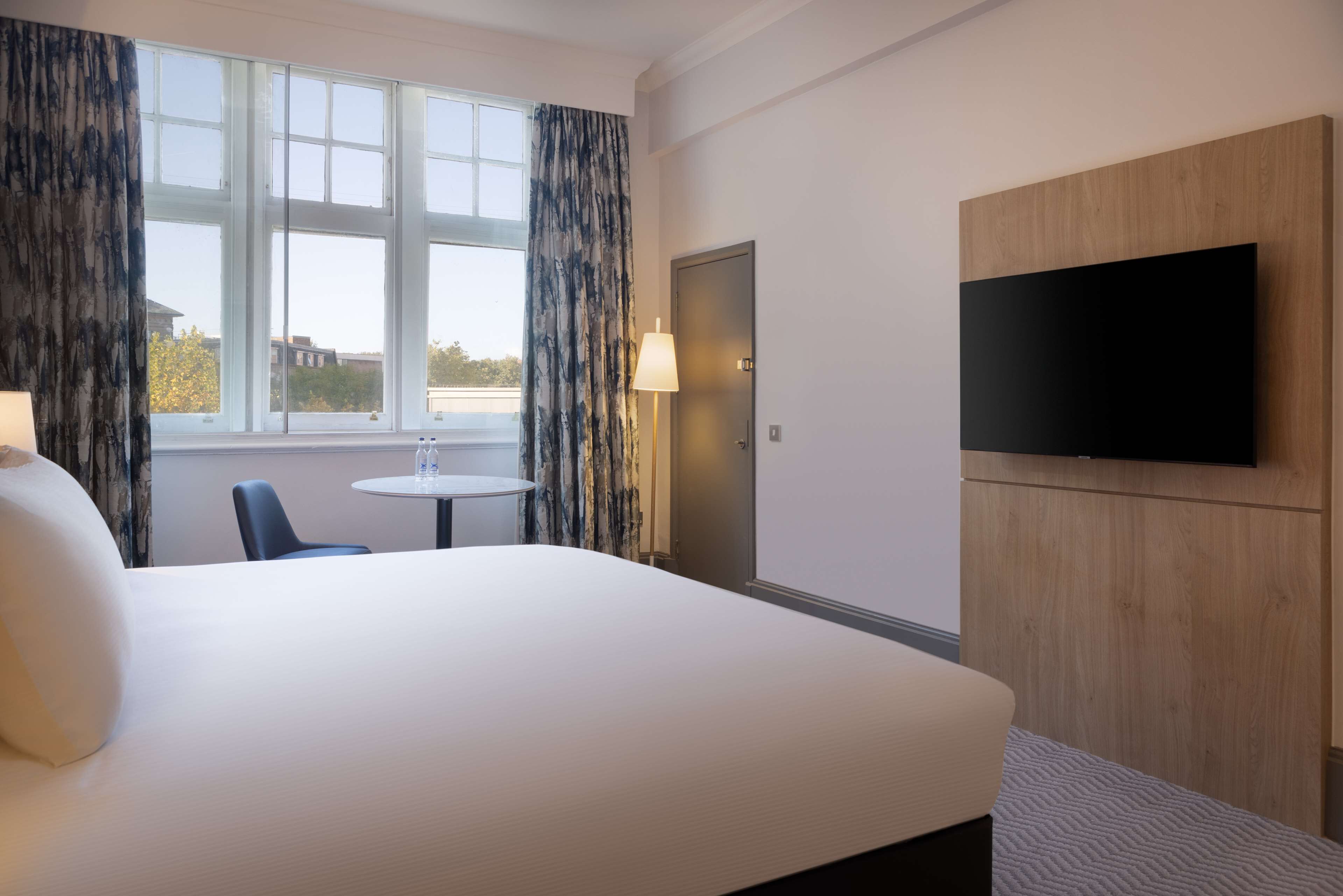 Une chambre d’hôtel avec un grand lit, un téléviseur à écran plat fixé au mur, une petite table ronde avec une chaise et une fenêtre avec des rideaux.