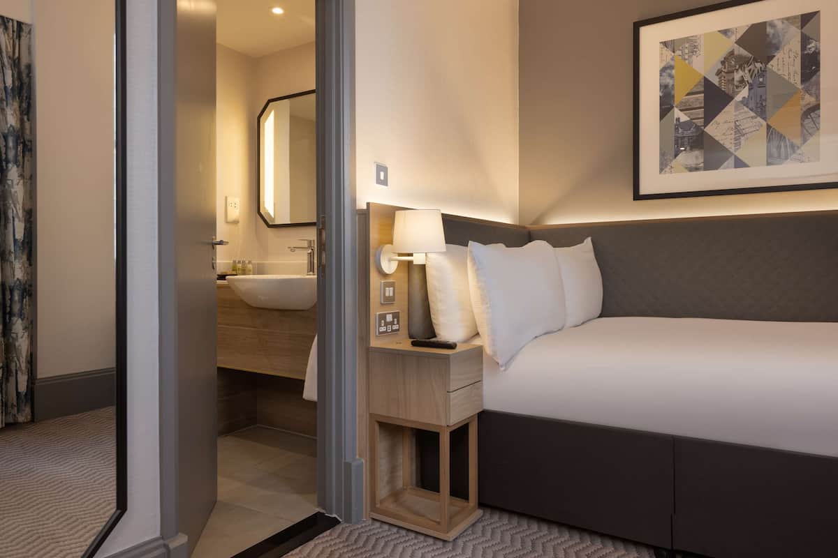 A hotel room with a bed, a bedside table with a lamp, a mirror, and a framed artwork on the wall.