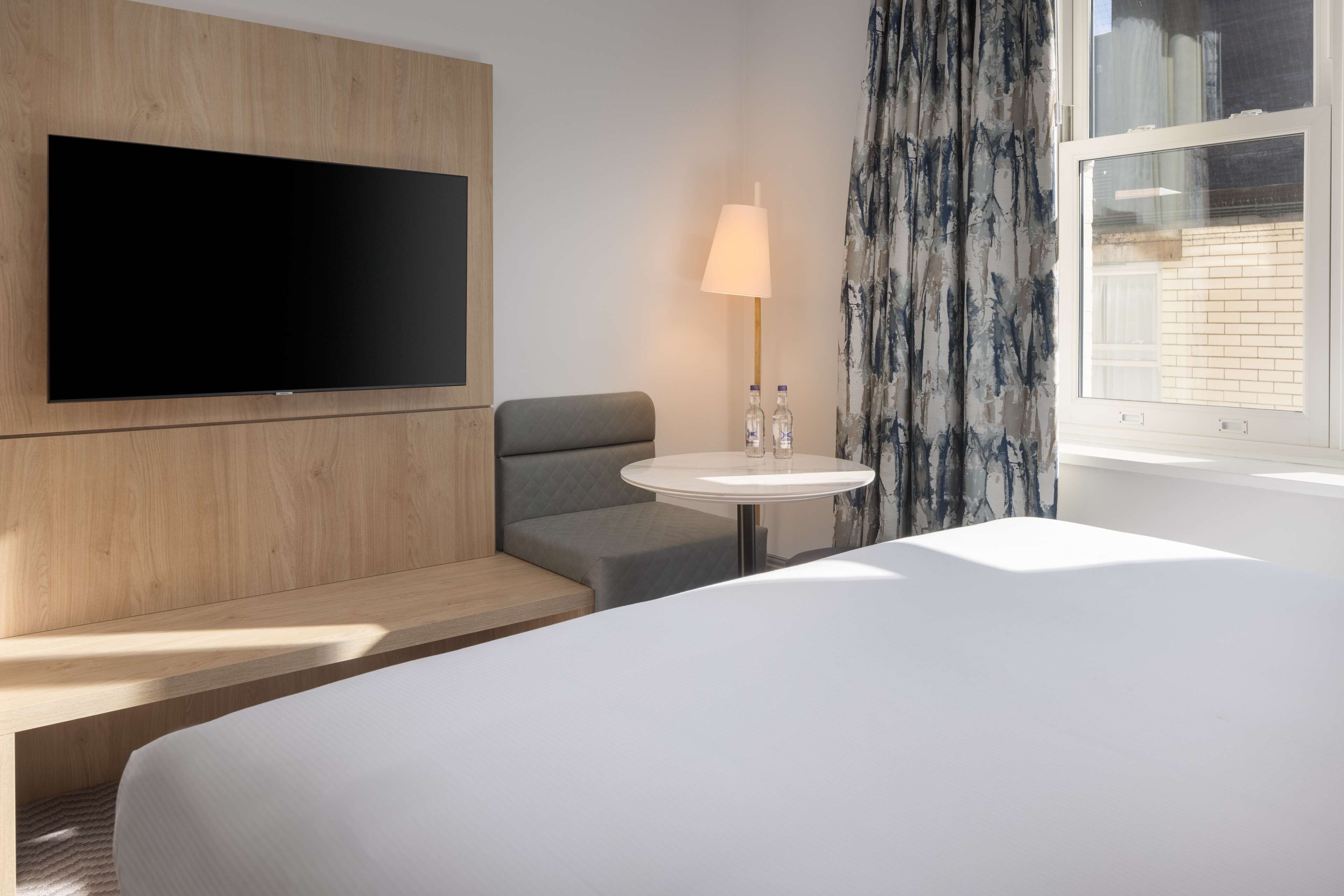 Moderni hotellihuone, jossa on seinälle asennettu taulutelevisio, harmaa sohva, pyöreä pöytä lampun kanssa ja suuri sänky.