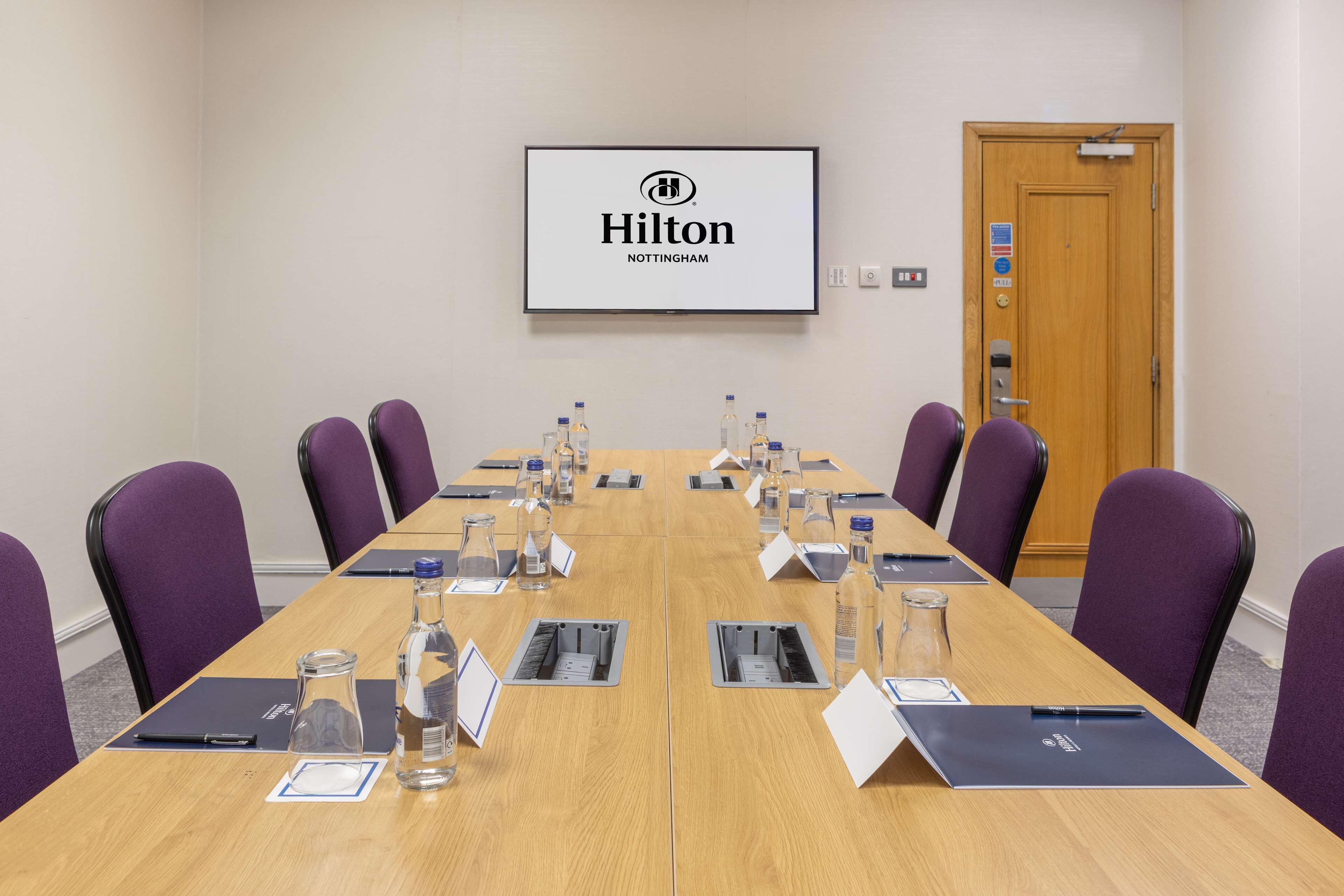 A conference room with a long table, chairs, name cards, water bottles, and a screen displaying the Hilton logo.