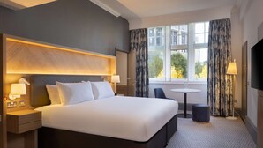 Habitación de hotel con una cama grande, mesitas de noche con lámparas, una pequeña mesa redonda y vistas al exterior a través de una ventana con cortinas.
