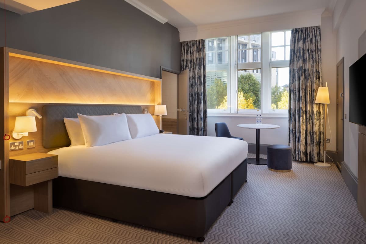 A hotel room with a large bed, bedside tables with lamps, a small round table, and a view of the outside through a window with curtains.