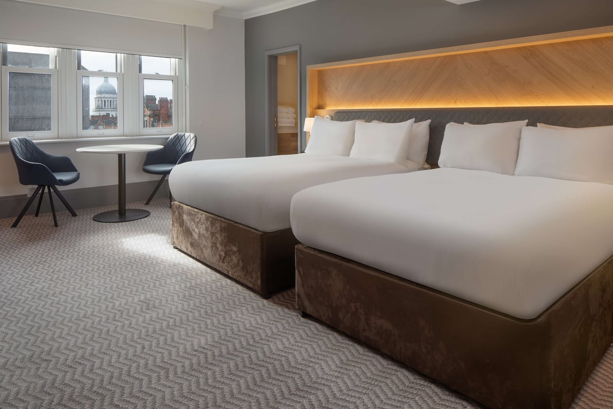 A hotel room with two beds, a small table, two chairs, and a view of a building through large windows.