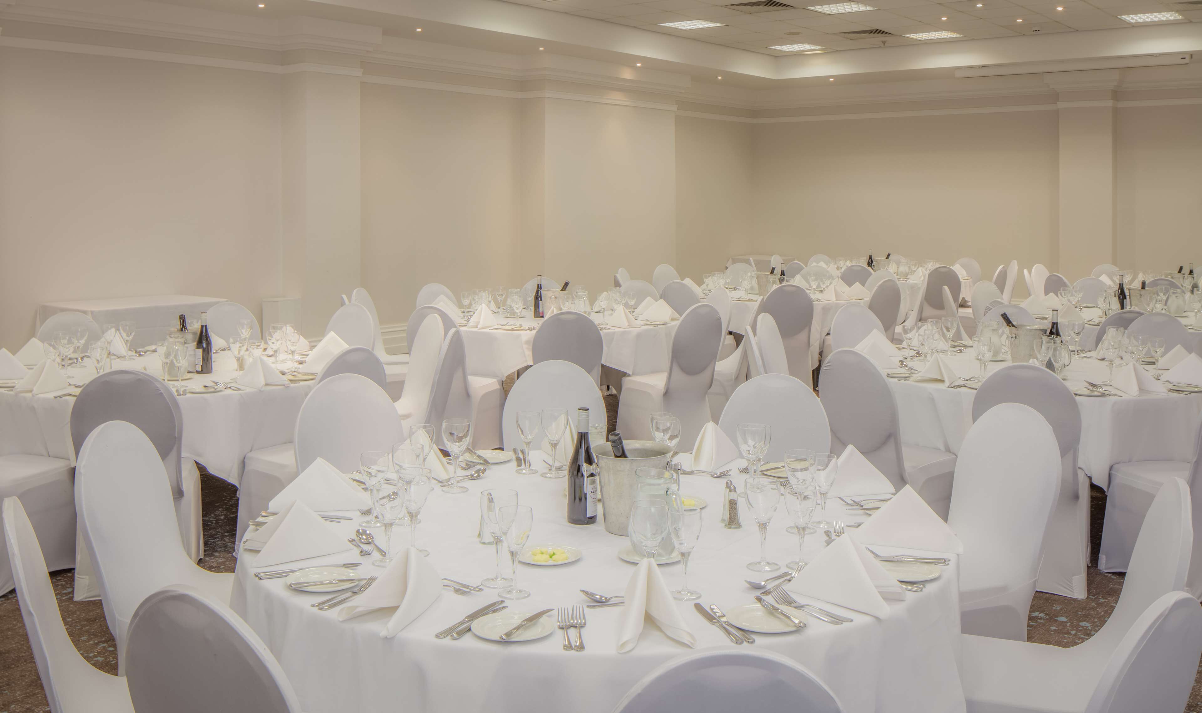 Un salón de banquetes con mesas redondas dispuestas para comer, sillas blancas y una paleta de colores neutros.