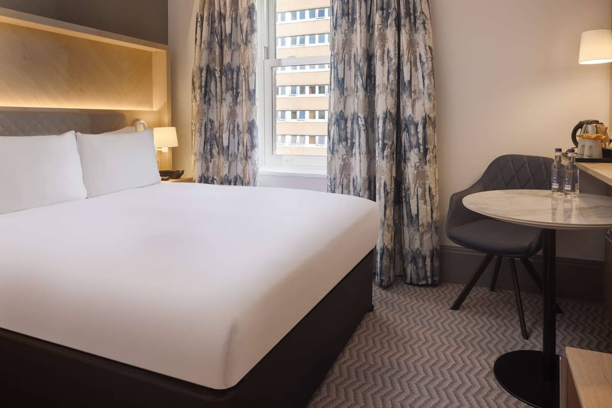 A hotel room with a large bed, a desk with a chair, a lamp, and a window with curtains.