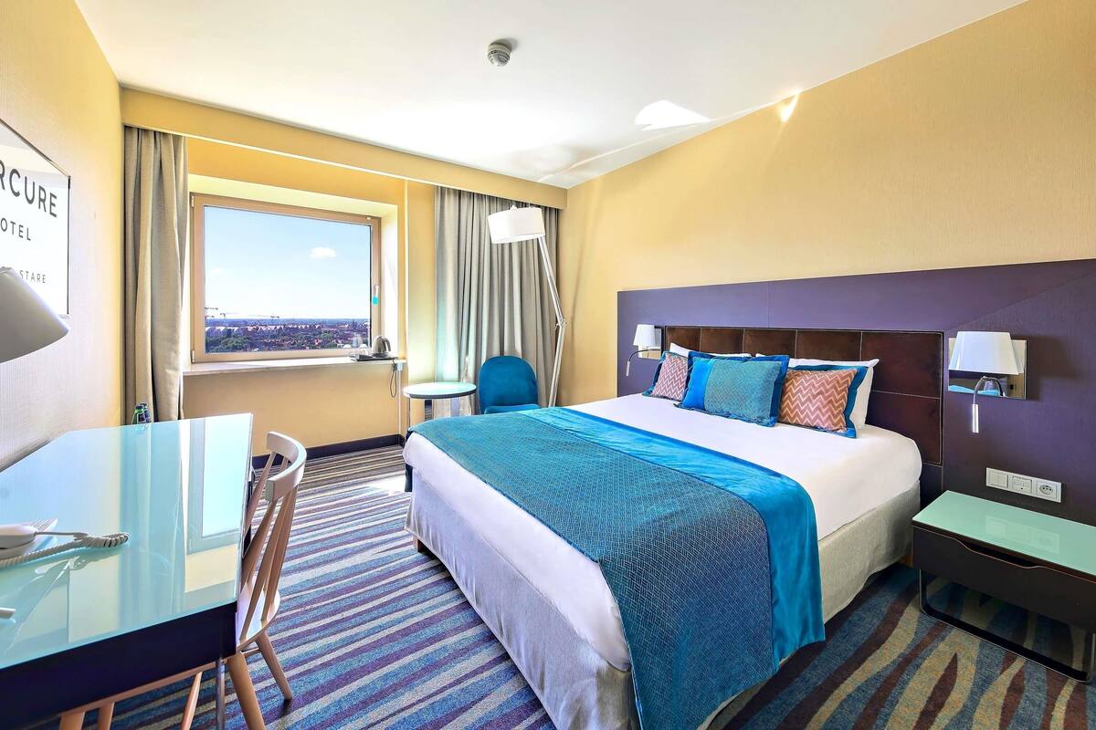Habitación de hotel con cama, escritorio, silla, ventana con vistas y detector de humo instalado en el techo.