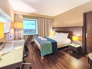 Una camera d'albergo con un letto, comodini, una scrivania con una lampada, una sedia, una finestra vista e una porta.