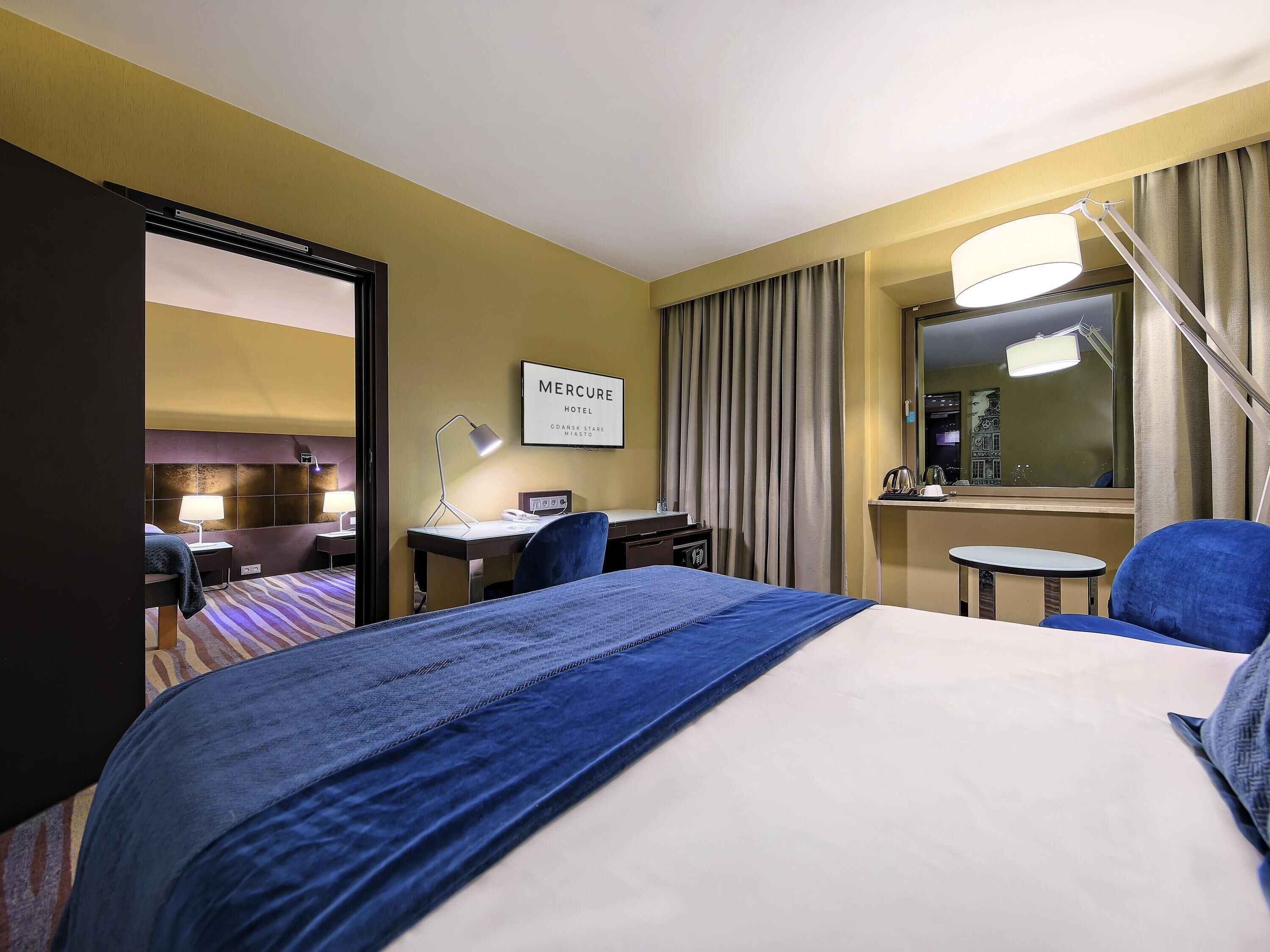 Habitación de hotel con cama, escritorio, silla, lámpara y un cartel que dice 'Mercure Hotel'.