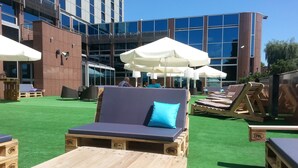 Un espace de détente aménagé sur le toit, avec des chaises longues en bois, des parasols blancs et une vue sur un bâtiment moderne aux fenêtres en verre.