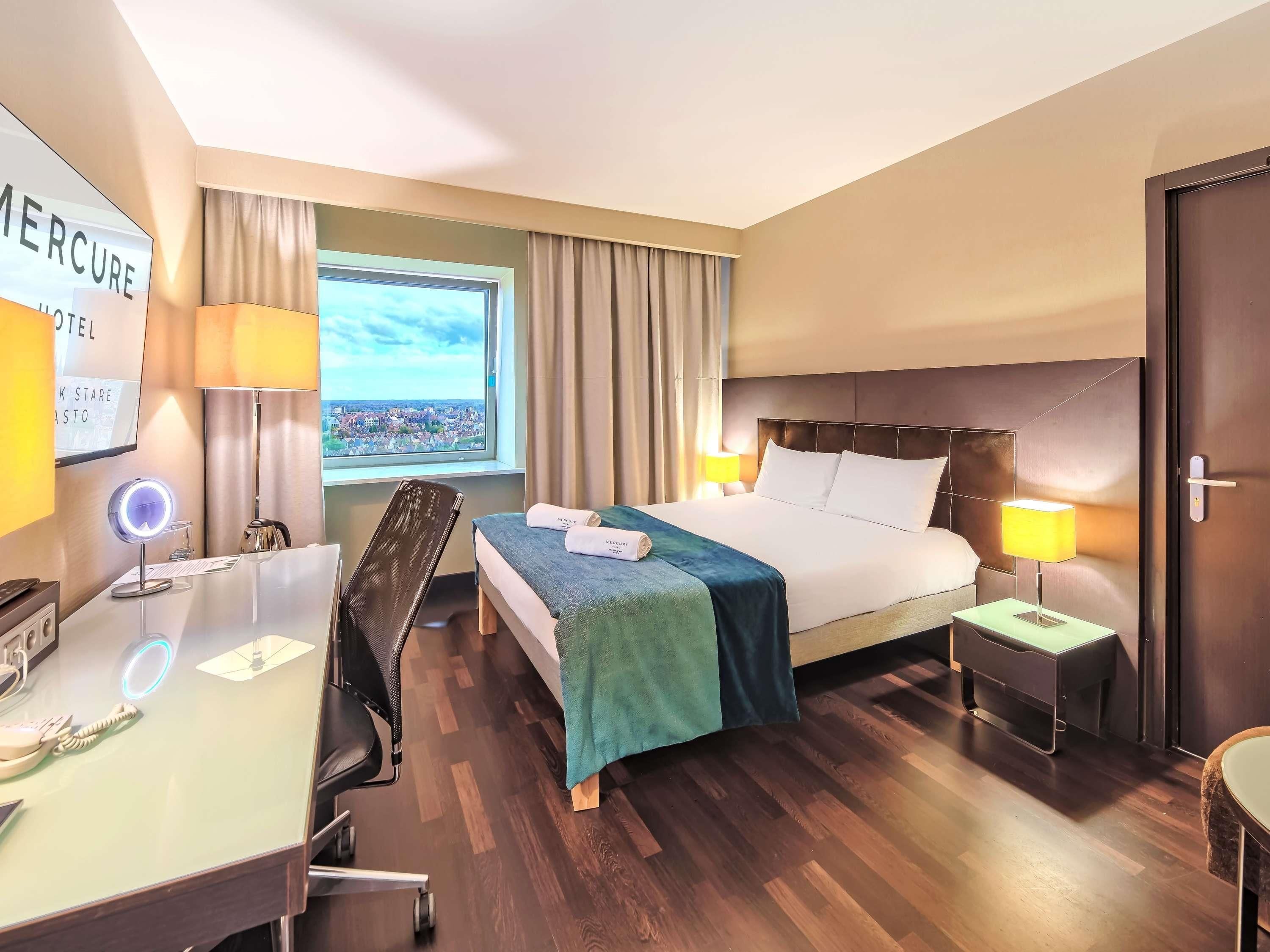 A hotel room with a bed, bedside lamps, a desk with a mirror, a chair, a small table, and a view of a cityscape through a window.
