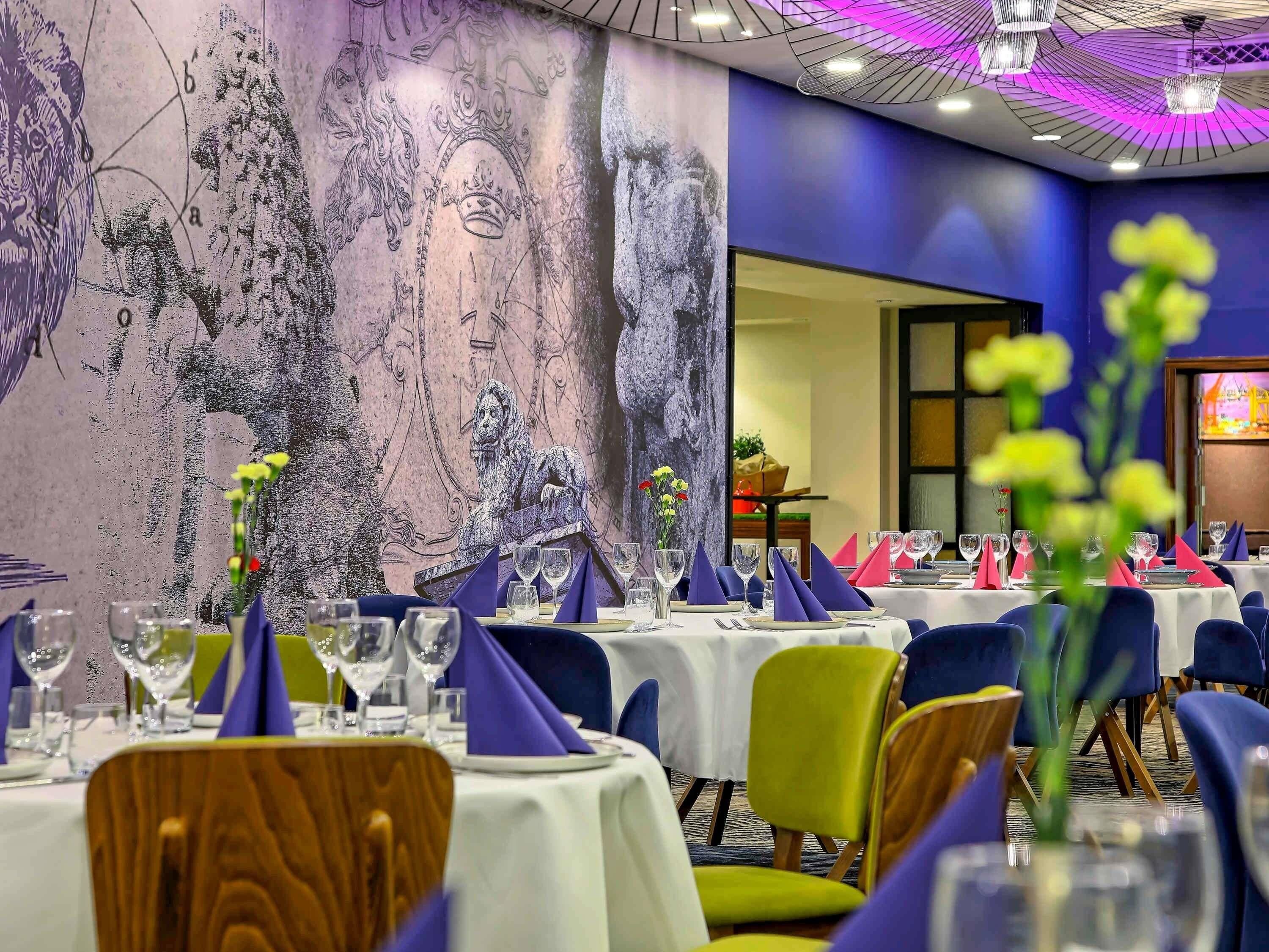 A banquet hall with round tables set for dining, featuring purple and green chairs, white tablecloths, and glassware.