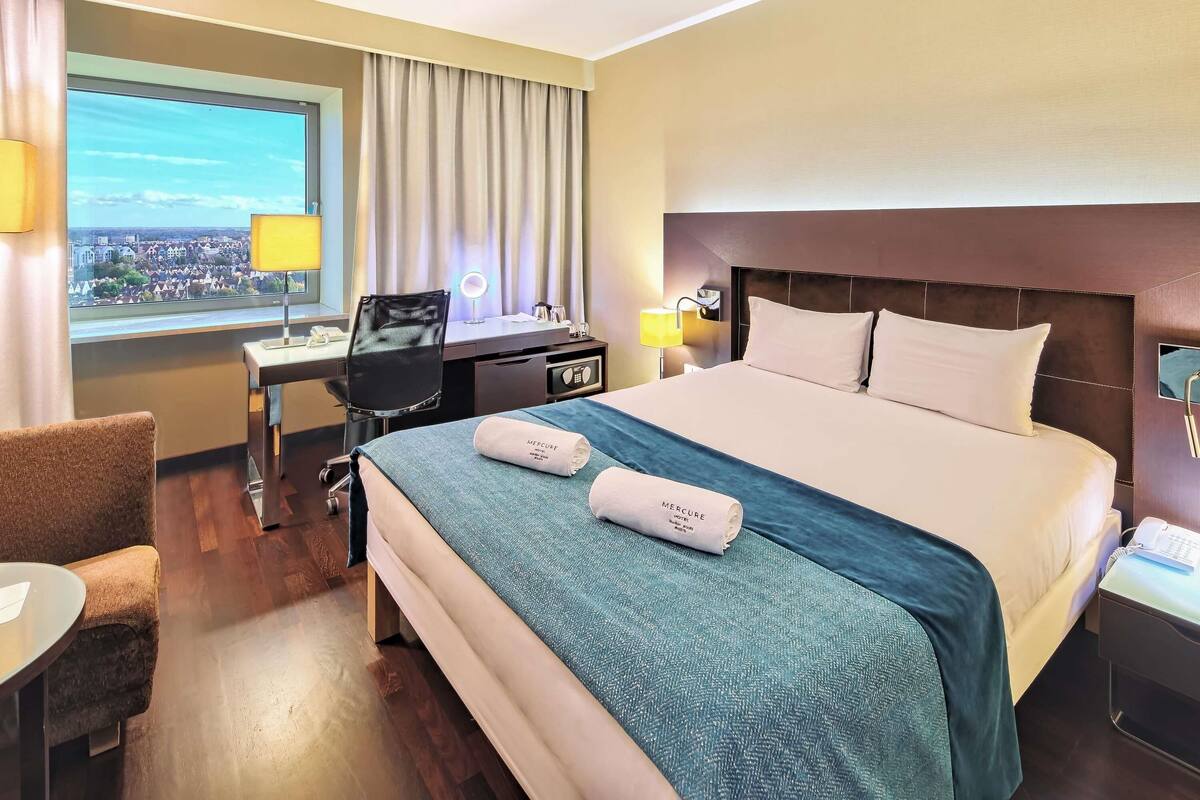 Habitación de hotel con una cama grande, un escritorio con una computadora, una silla, una ventana con vista a un paisaje urbano y una mesita de noche con lámpara.