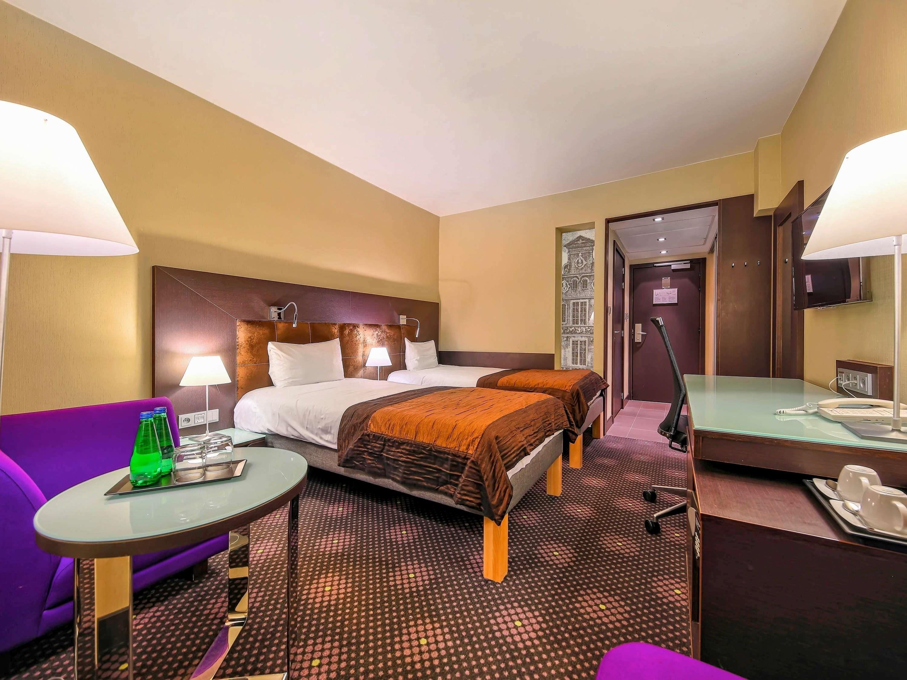 Une chambre d’hôtel avec deux lits, un canapé violet, une petite table avec une lampe, un bureau avec un téléphone et un miroir.
