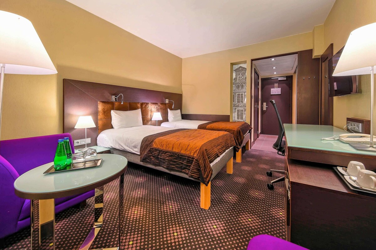 A hotel room with two beds, a purple sofa, a small table with a lamp, a desk with a telephone, and a mirror.