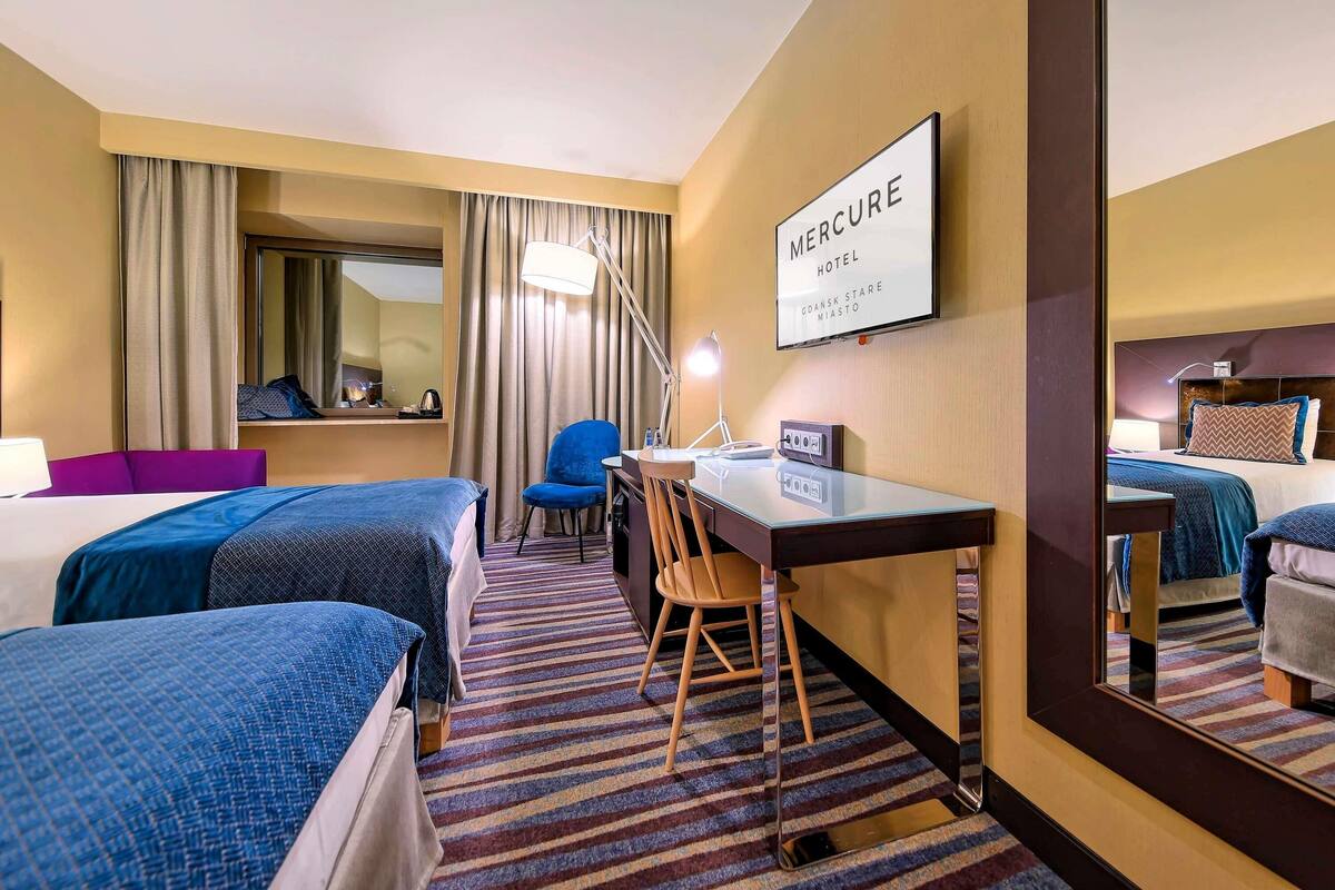 Habitación de hotel con dos camas, un escritorio, una silla, un espejo y un cartel con el nombre del hotel.