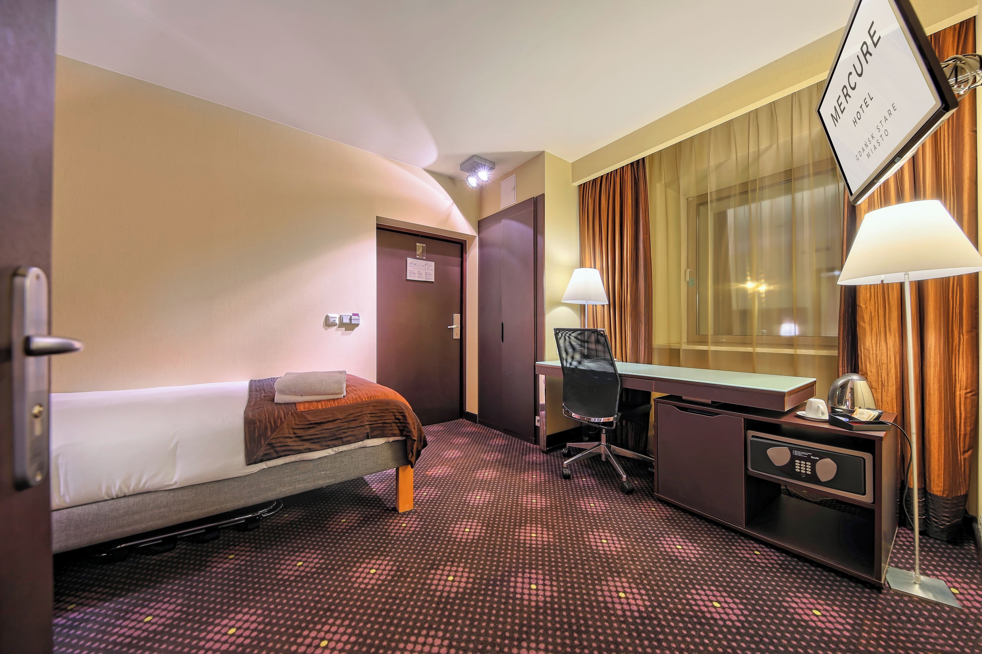 A hotel room with a bed, desk, chair, and a sign indicating it's the Mercure Hotel.
