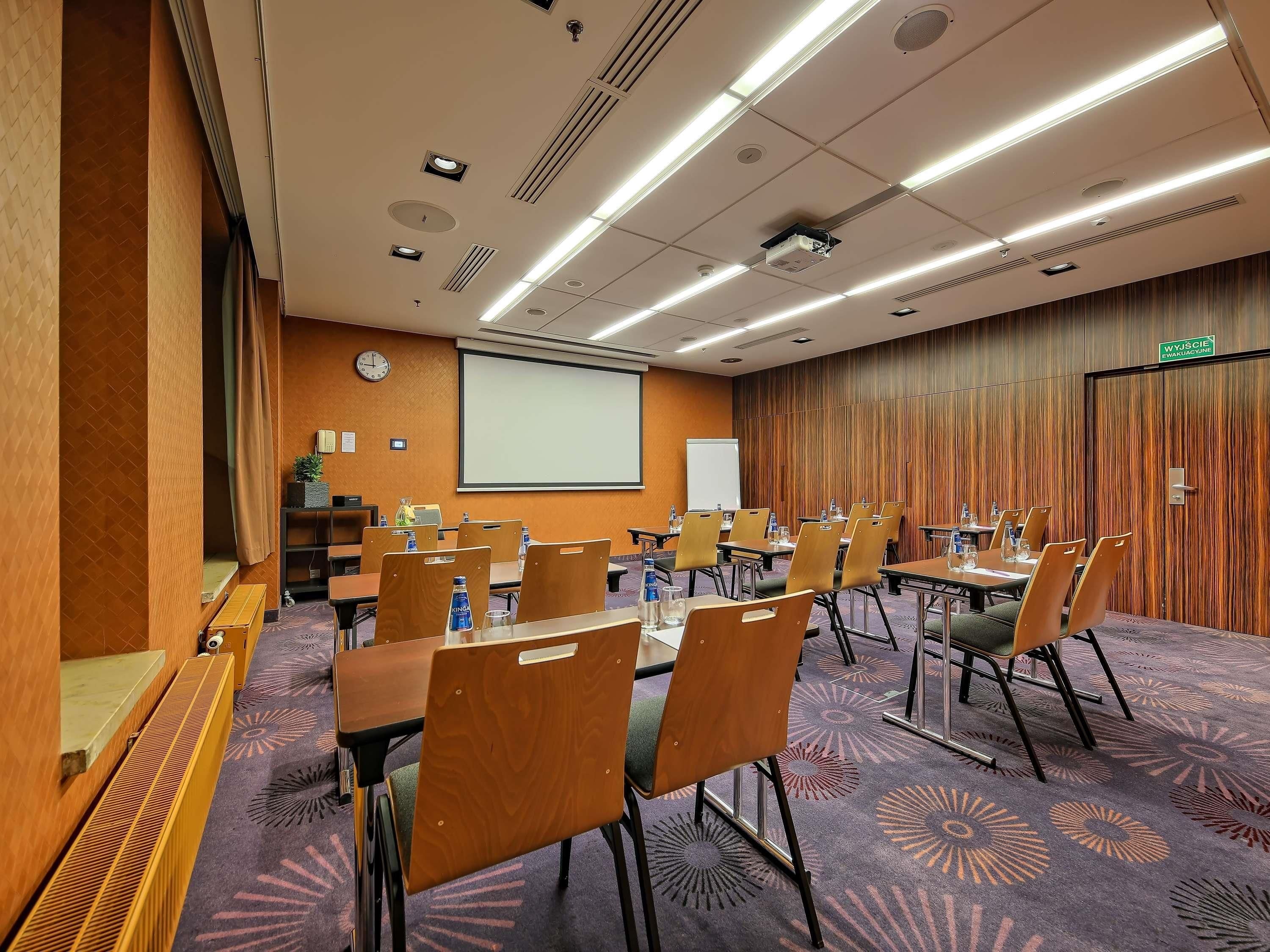Een vergaderzaal met een beamer, scherm en meerdere tafels met stoelen.