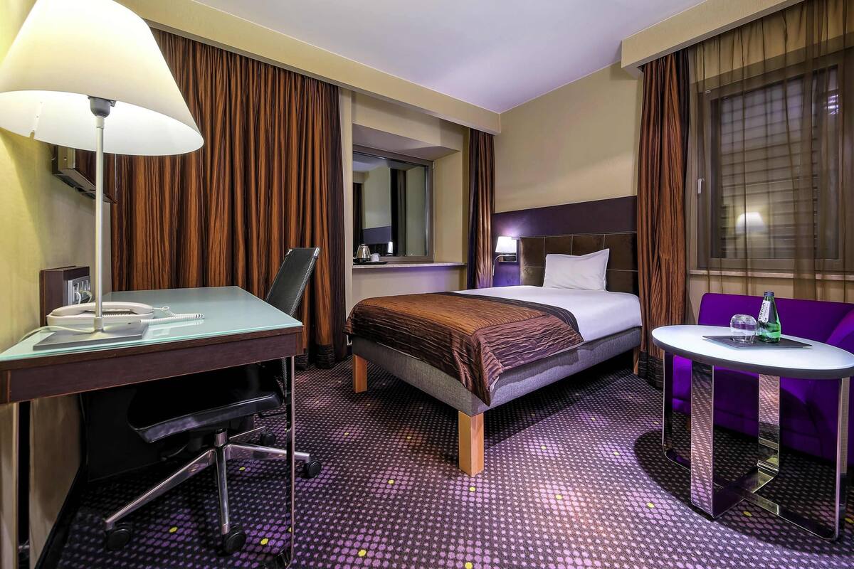 Habitación de hotel con cama, escritorio, silla y una mesita con una botella y un vaso.