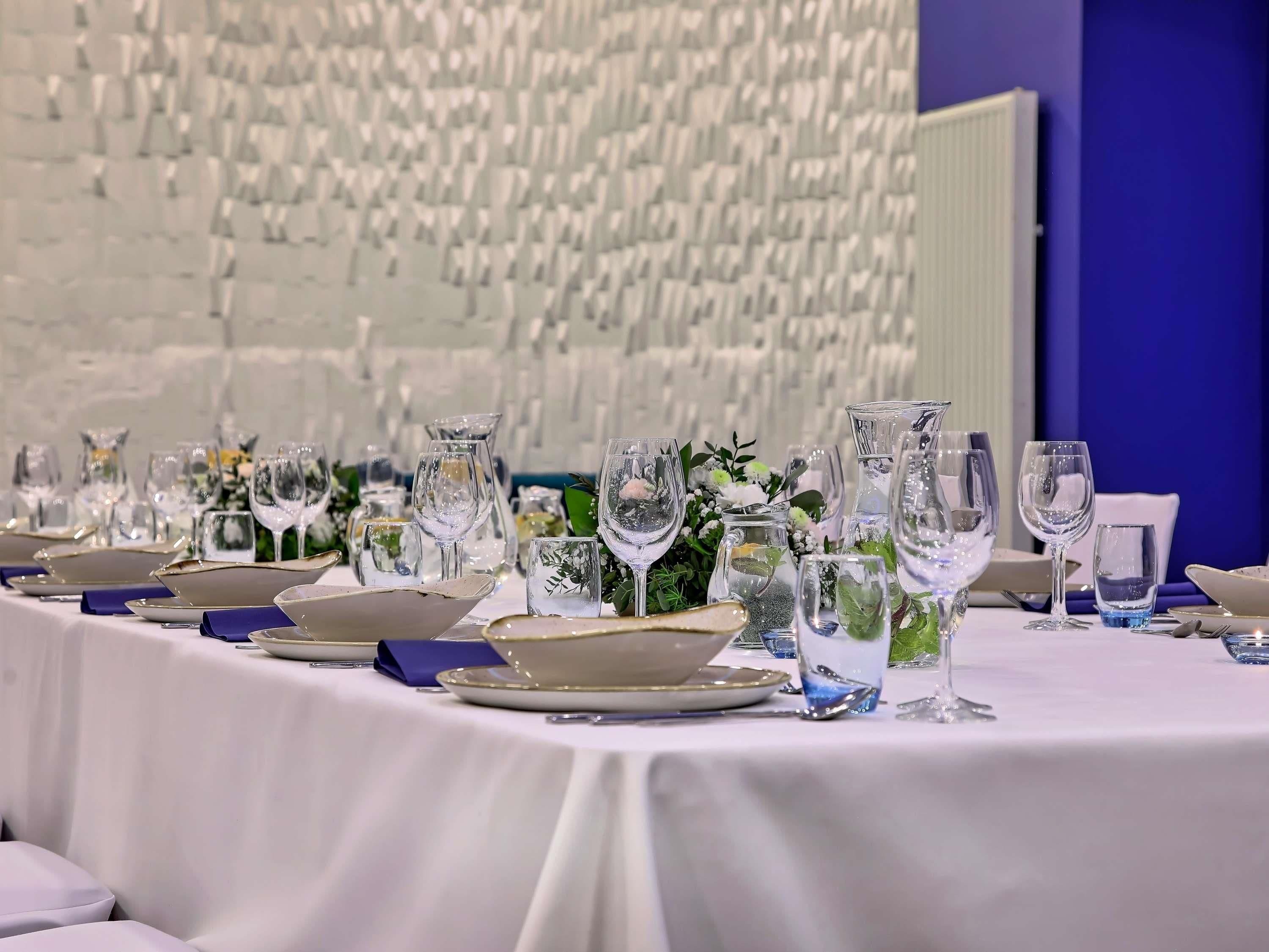 A table set with clear glassware, plates, and blue napkins, with a white tablecloth and a decorative wall in the background.