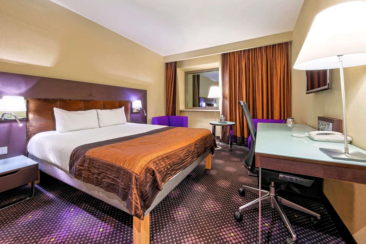 Une chambre d’hôtel comprenant un lit, un bureau, une chaise, une petite table, une lampe, un téléphone et un miroir.