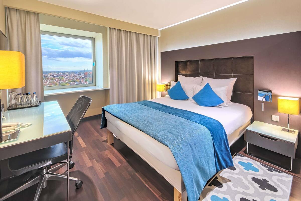 Habitación de hotel con una cama grande, un escritorio, una silla, una lámpara de escritorio, una mesita de noche, una lámpara, vistas a un paisaje urbano y una alfombra estampada.